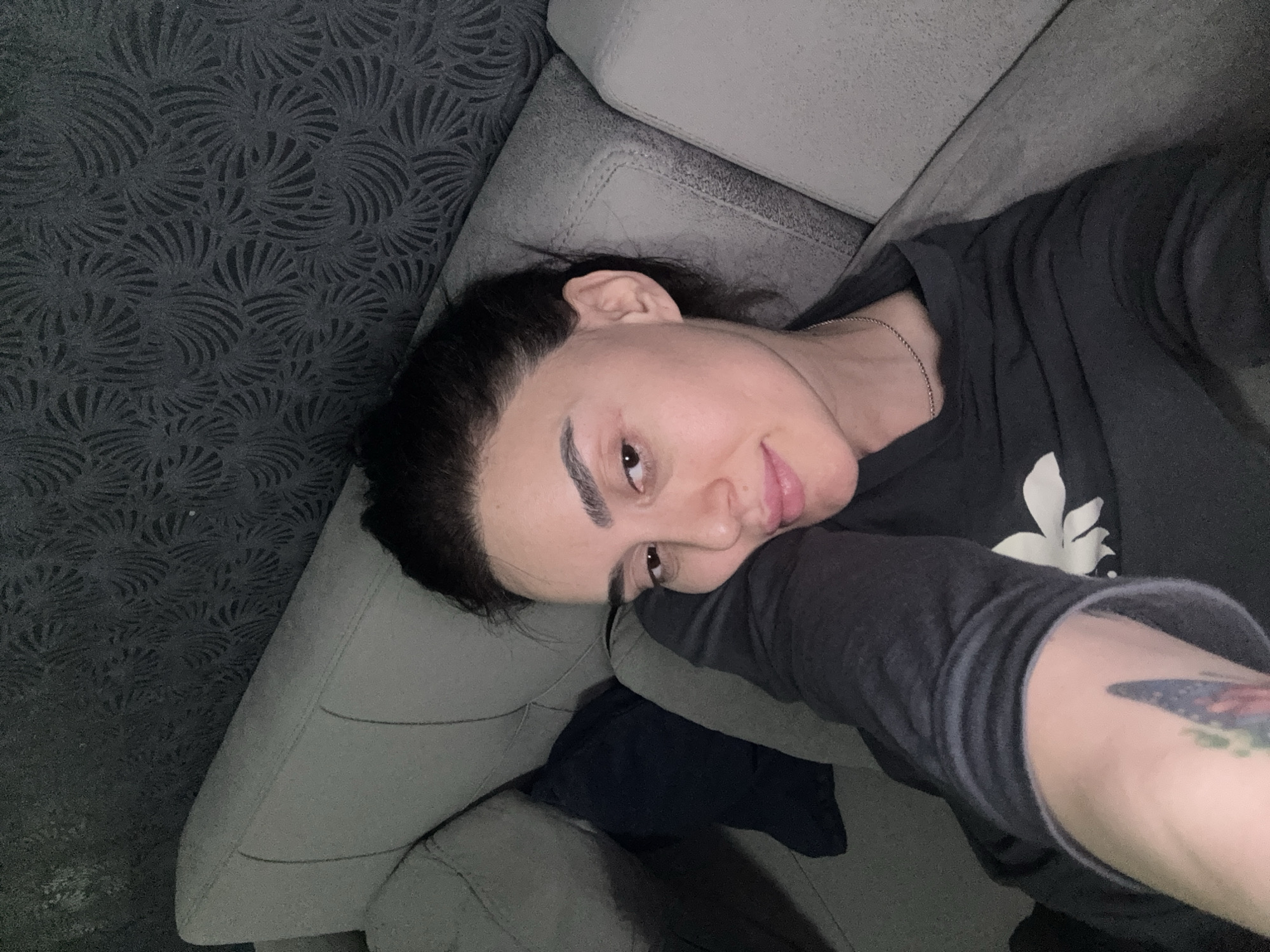A person is lying on a gray couch. They have dark hair pulled back from their face, prominent eyebrows, and a slight smile. They are wearing a dark-colored, long-sleeved shirt with a white floral design. The person's right arm is visible, bearing a tattoo. The couch is against a wall with a dark, patterned wallpaper. The location cannot be identified as Tbilisi, Georgia based on the information present in the image.