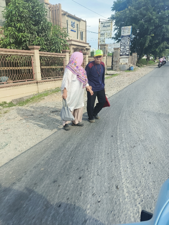 Two people, a woman and a man, are walking in the street in Sukatani, Indonesia. The woman is wearing a pink hijab and a white dress, carrying a grey bag. She is walking on the left side of the street. The man is wearing a blue shirt, grey pants, and a yellow hat. He is walking on the right side of the street. They are walking in the direction of a blue car that is in the foreground of the image. The street is paved with asphalt. There is a building on the left side of the street with a sign that reads "Aurosal". A black metal fence separates the building and the street. There are trees lining both sides of the street. The weather is sunny. The GPS coordinates are -6.623 latitude, 107.404 longitude.