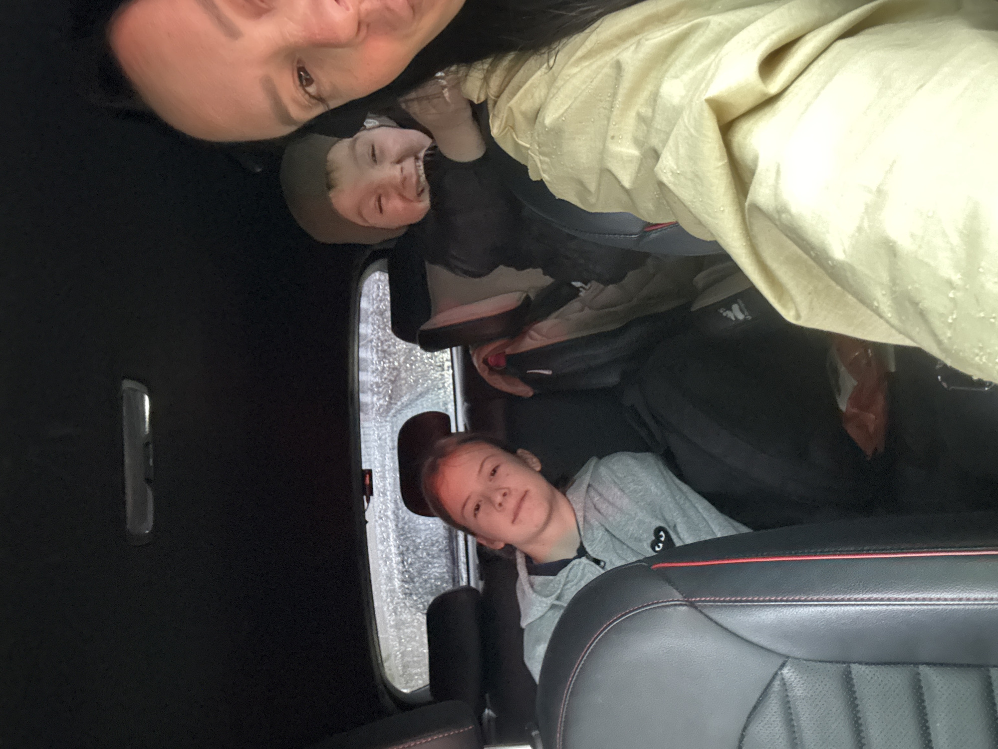 A selfie inside a car shows a woman and two children. The woman is at the top of the frame, and the child is in a car seat covered with a yellow coat. The other child is in the backseat wearing a gray sweatshirt. The car window shows rain. The location is Chaikovsky, Russia.