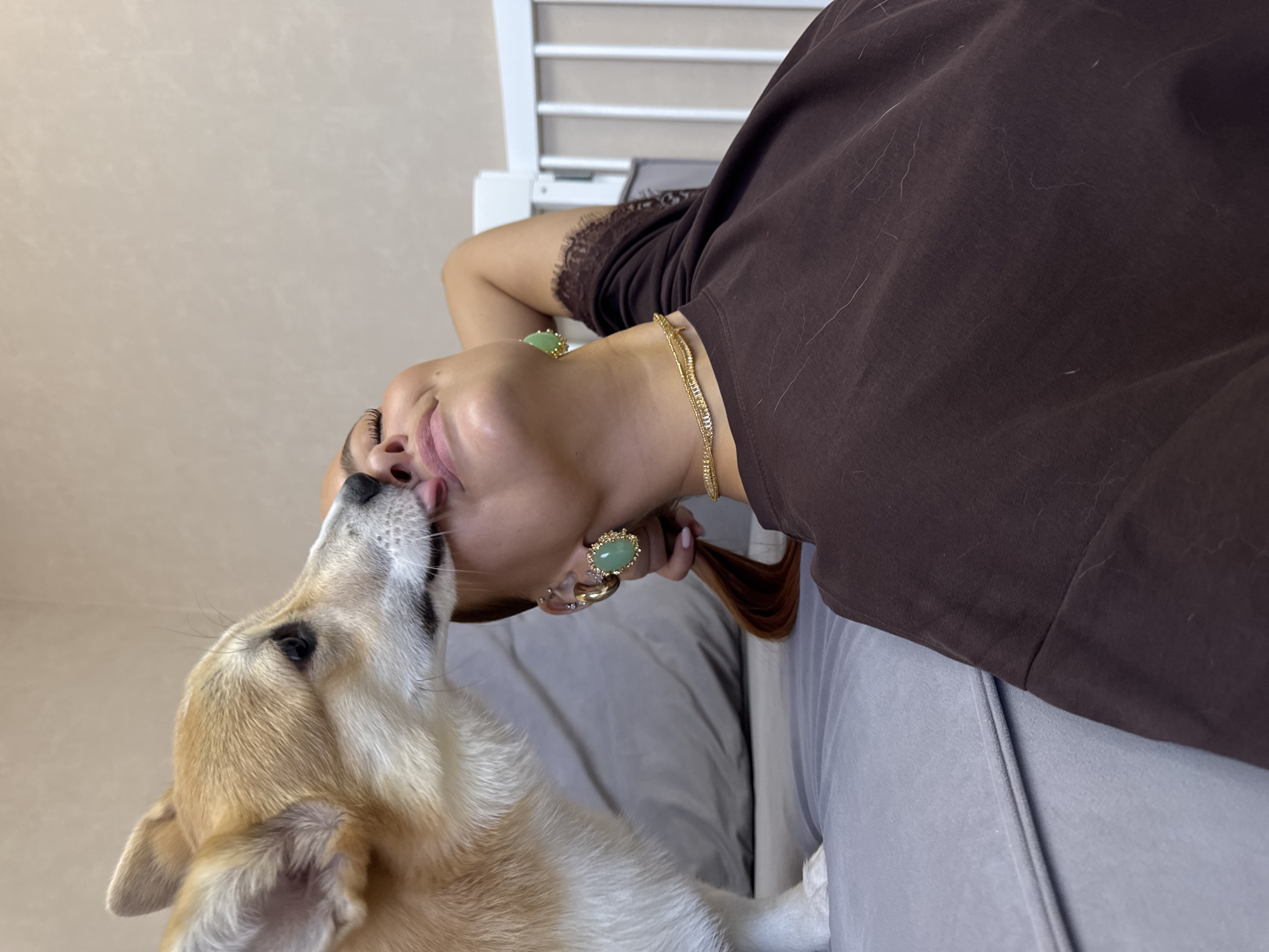 A fair-skinned woman is lying on her back on light gray bedding, with a tan and white dog standing near her face, licking her lips. The woman is wearing a dark brown, short-sleeved top with brown lace detail on the sleeve and gray pants. She is also wearing gold earrings with a green stone, a gold bracelet, and other gold jewelry on her ears. The background includes an off-white wall and a white structure, possibly a towel rack or furniture, near the top of the image. The photo appears to have been taken from a low angle. The image does not contain any evidence to suggest it was taken in Chaikovsky, Russia.