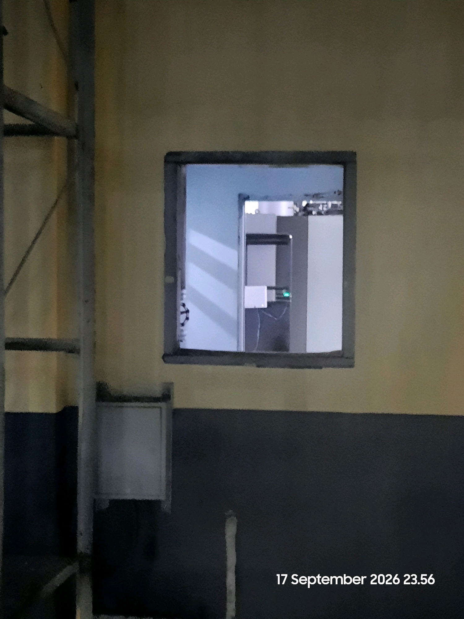 This is a photograph of a building exterior with a small window. The wall is painted in two horizontal sections: the upper section is a pale yellow, and the lower section is a dark gray. A metal ladder or scaffolding is visible on the left side of the image. There is a small, square metal box mounted on the dark gray section of the wall below the window. The window itself is rectangular and appears to look into an interior space. The interior appears to have a light blue-gray wall with some shelving units and what looks like a white box with a green light on it. The overall lighting suggests it is nighttime. The date and time "17 September 2026 23.56" are superimposed on the bottom right of the image. The location is identified as Gending, Indonesia.