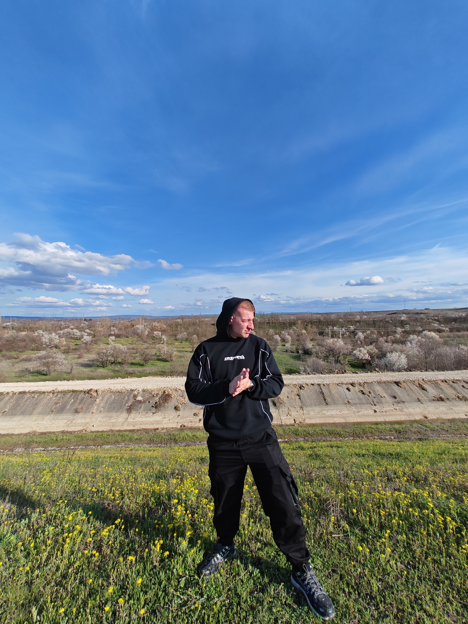 A man stands in a field of yellow flowers, with a concrete drainage channel and flowering trees in the background, under a blue sky with scattered clouds. The man is wearing a black hooded sweatshirt with white trim, black pants, and sneakers. He is facing left, with his hands clasped in front of him. The landscape suggests the outskirts of a developed area, possibly in the vicinity of Shumen, Bulgaria.