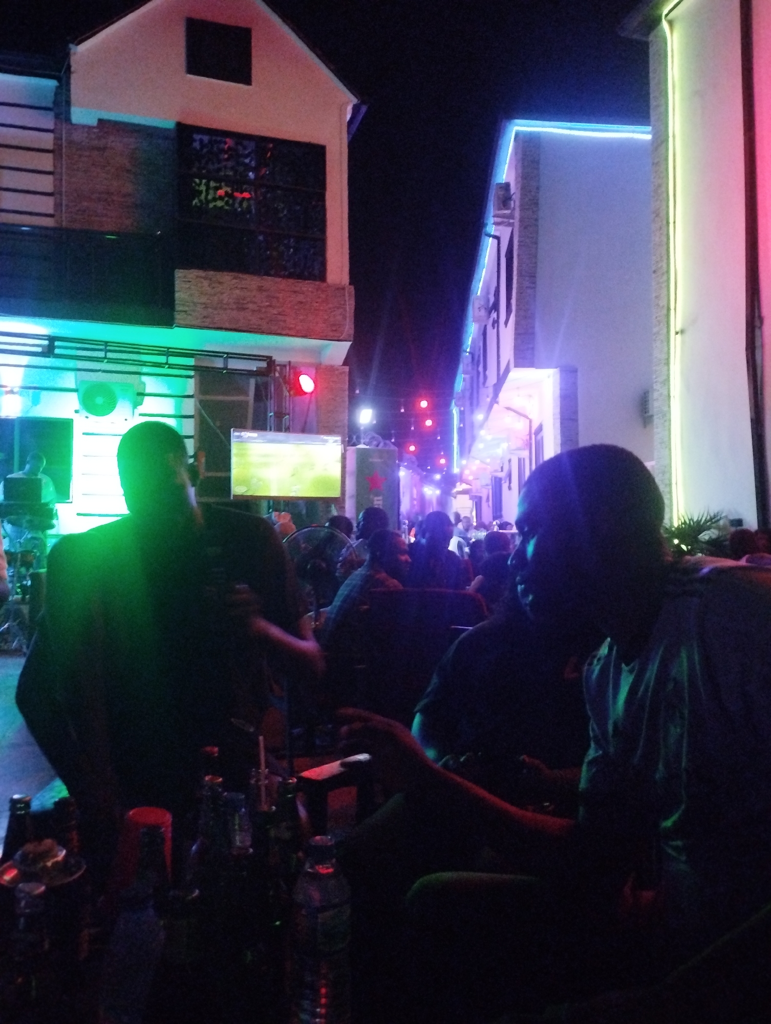 The image shows a nighttime scene at what appears to be an outdoor bar or lounge in Tumfure, Nigeria. Several people are seated at tables. At least two buildings are visible in the background, one with a brick facade and another with white exterior walls. The scene is illuminated by colorful neon lights (green, blue, pink). There is a TV screen showing what appears to be a soccer game. Several bottles are on the table in the foreground. The overall setting seems to be a gathering or social event in the city.
