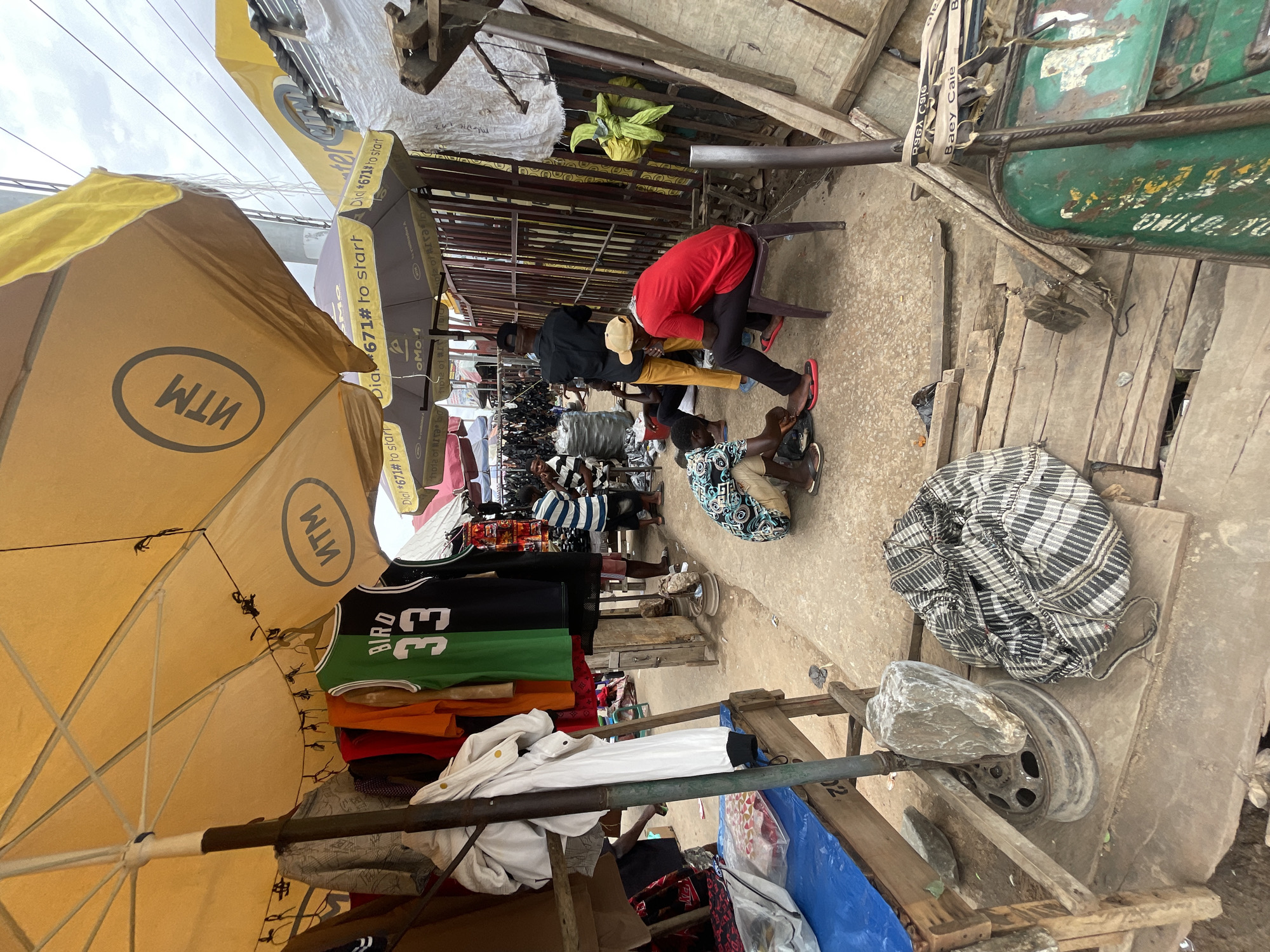 The image depicts a street scene, likely a marketplace in Onitsha, Nigeria, based on the presence of MTN-branded umbrellas, a common sight in West African markets. People are clustered together in the middle of the frame. One person in a red shirt is seated on a small chair. Another person in a black jacket and khaki pants stands close behind. In front, another person is kneeling on the ground. This group appears to be engaged in some sort of task. Clothing items including a "Bird 33" jersey are hanging for sale under the umbrellas. Other merchants and potential customers can be seen in the background. A makeshift cart with a large stone rests in the foreground. The environment is densely populated, with various goods and structures crowding the space.