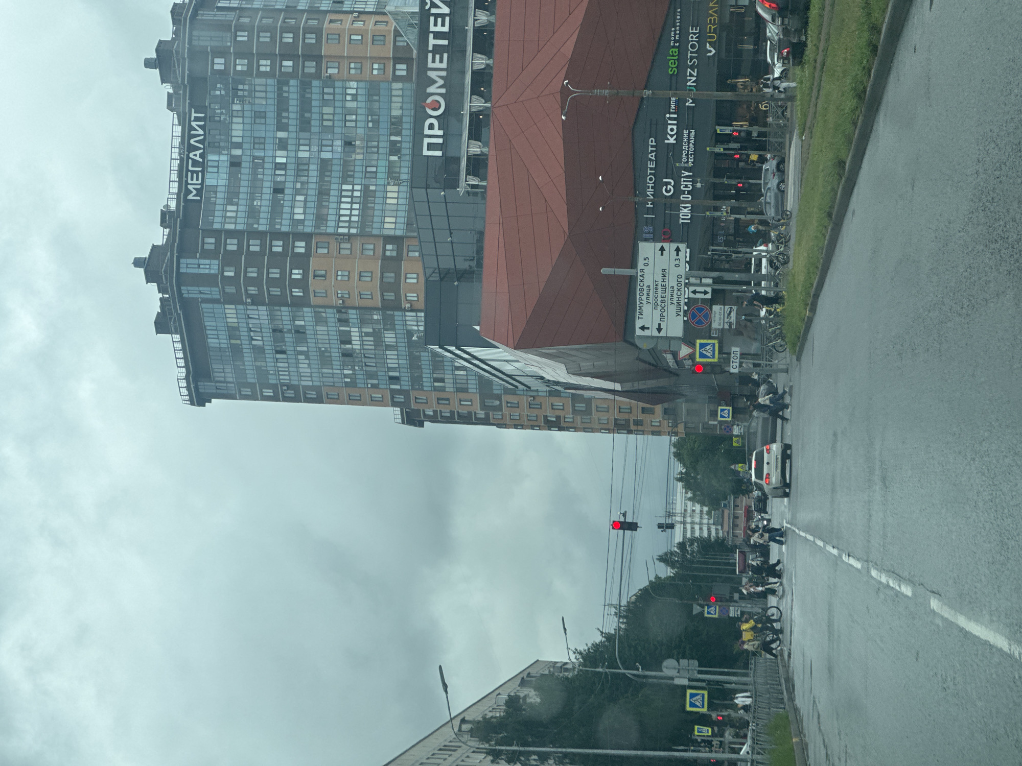The image depicts a city street scene in Saint Petersburg, Russia, on a cloudy day. A tall, modern residential building with a dark facade and numerous windows dominates the right side of the frame. The building has "МЕГАЛИТ" (MEGALIT) written in large white letters near its top. Below this building, a commercial complex with a distinctive red, angular roof structure is visible, featuring a sign for "ПРОМЕТЕЙ" (PROMETEY) in white letters. To the left of the residential building, a lower, light-colored apartment building stretches along the street.
The street in the foreground is wet, suggesting recent rain. Several cars are on the road, and pedestrians are crossing at a crosswalk. Traffic lights are visible, with one showing a red signal. There are also various street signs and traffic signals. In the background, more buildings and trees can be seen. The overall atmosphere is urban and overcast.