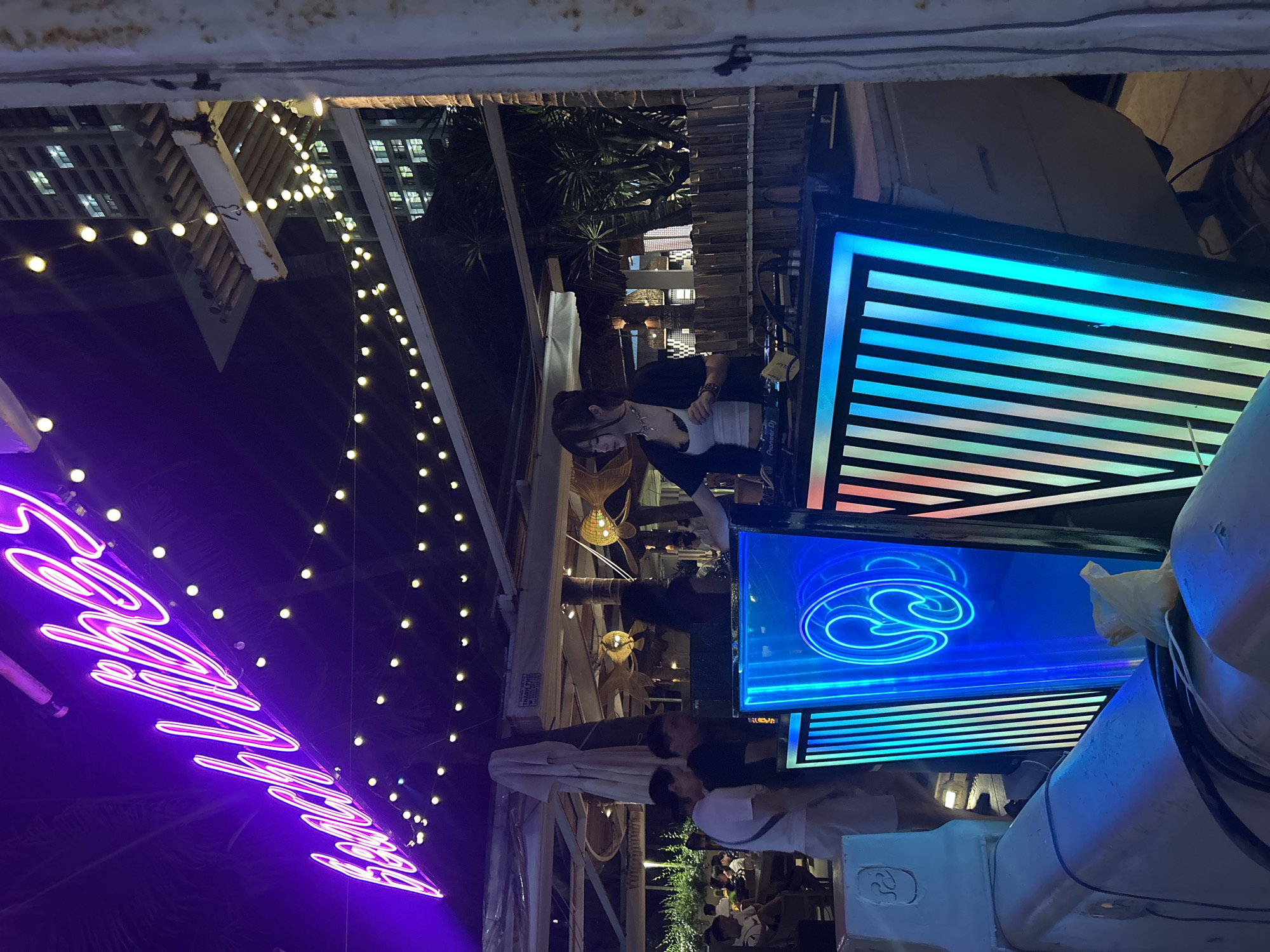 This image captures a vibrant outdoor or semi-outdoor venue, likely a bar or club, at night. The scene is illuminated by numerous decorative string lights with glowing white bulbs, creating a festive ambiance.
A female DJ is positioned at the center-right, standing behind a DJ booth. She has dark hair tied back and is wearing a light-colored top under a dark jacket, focused on her "Pioneer DJ" equipment. Behind her, two large screens are prominent features of the DJ setup. The upper screen displays horizontal stripes in shades of blue, while the lower screen features a glowing, intricate blue swirling graphic or logo.
To the left, a large, purple neon sign spells out "Good Vibes," contributing to the lively atmosphere. Several patrons are visible in the background, seated at tables or standing, enjoying the evening. Architectural elements, including wooden beams and structures, are noticeable, along with decorative hanging lamps, some appearing to be made of wicker. Palm trees in the background suggest a warm climate. A small, white plastic bag is also seen hanging near the right screen. The overall mood is energetic and inviting, indicative of an evening social event.