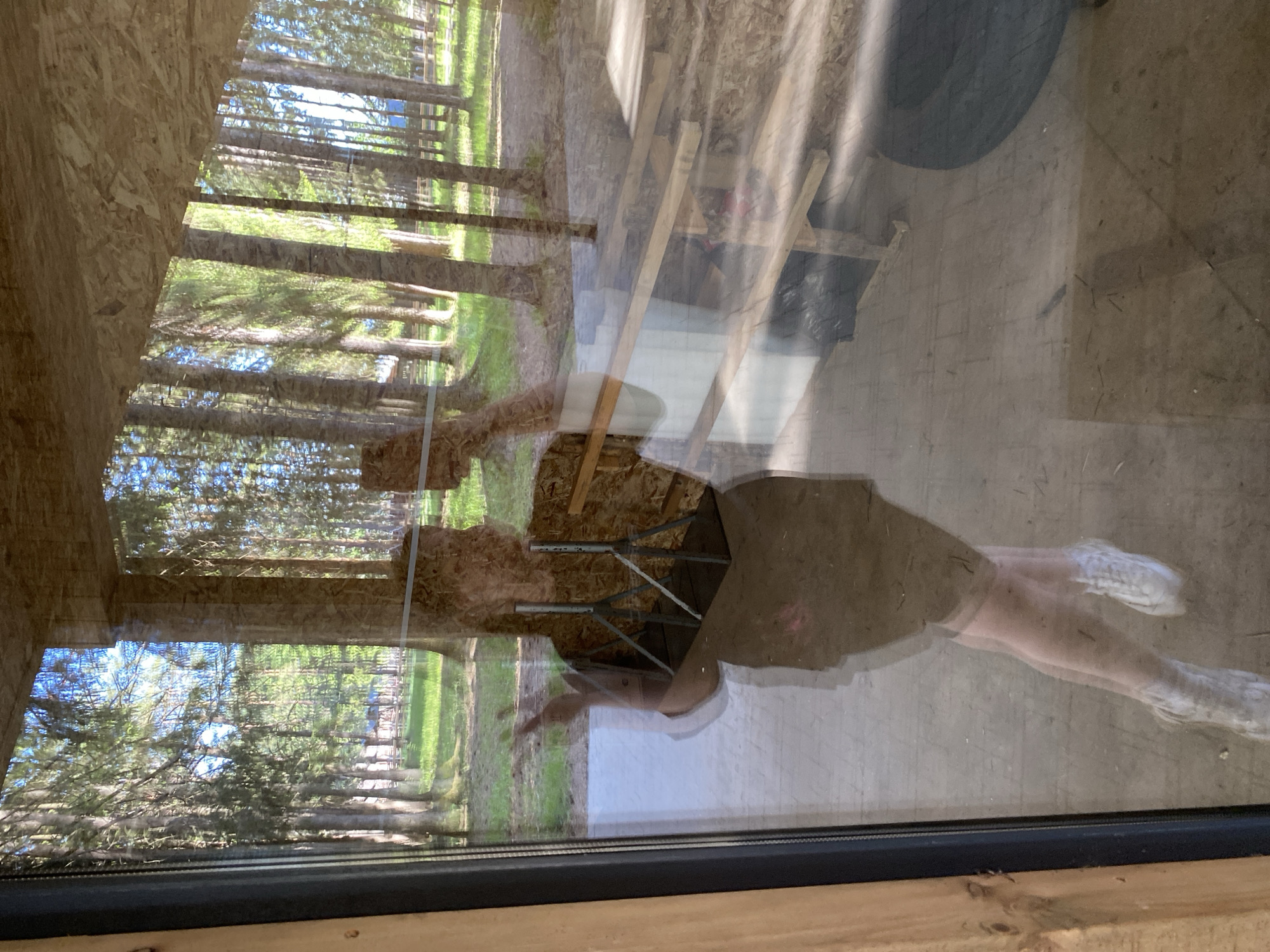 The image shows a reflection in a window. The reflection includes a person wearing white sneakers, shorts, and a t-shirt. Also reflected are trees, possibly a wooded area. The interior visible through the window includes building materials (wood, boards) and some construction equipment/materials. The location is unknown, with no clear indicators of being in Votkinsk, Russia.