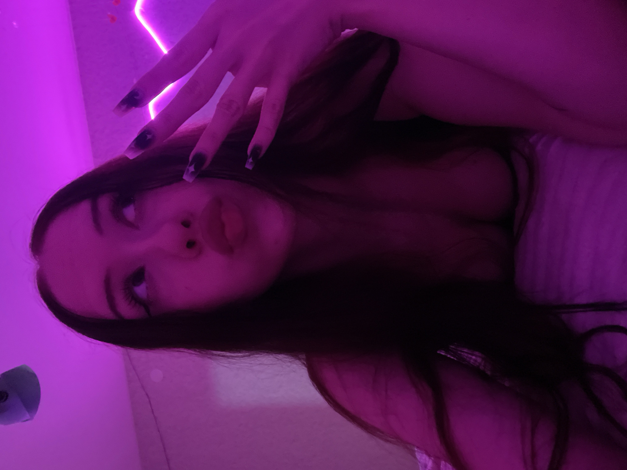 The image shows a close-up of a young woman with long dark hair, lying down and looking upwards. She is illuminated by pinkish-purple light, which casts a strong hue over the scene. Her lips are slightly parted, and her eyes are looking away from the camera. Her hand, with long dark fingernails, is raised near her face, obscuring part of her hair. The background appears to be a wall with a neon light strip, possibly a room interior. The overall impression is of a selfie taken in a dimly lit, perhaps stylized, environment. The location in Votkinsk, Russia is not determinable from the visual information provided in the image.
