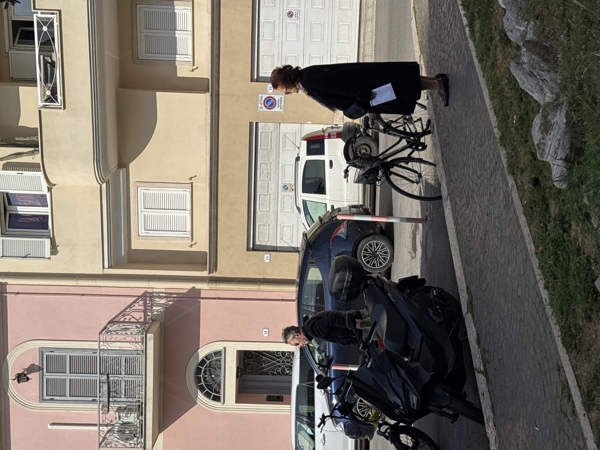 The scene shows a street in Nettuno, Italy, with buildings on the left side of the frame. Two people are visible; one is a senior woman in a dark coat walking with a bicycle, and the other is a man standing by a car. A dark blue car, a white van, and a motorcycle are parked along the street. Building façades are visible, with various architectural details like windows with shutters and decorative balconies. The street is paved, and there is a grassy area on the right side of the frame. The road sign with the number 15 indicates the number of the building.