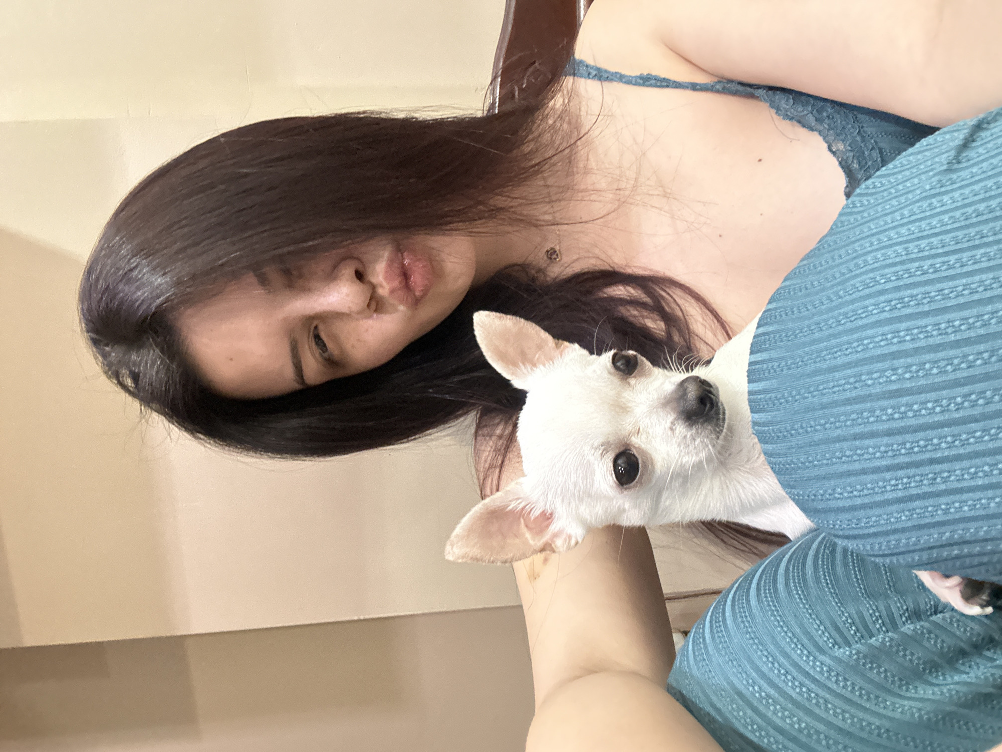 A woman with long, dark hair is lying down, making a kissing face towards the camera. She is wearing a blue lace top. A small, white Chihuahua is nestled in her arms, looking directly at the camera. The background consists of a beige wall and a wooden chair. The exact location within the city of Sliven, Bulgaria, cannot be determined from this image.