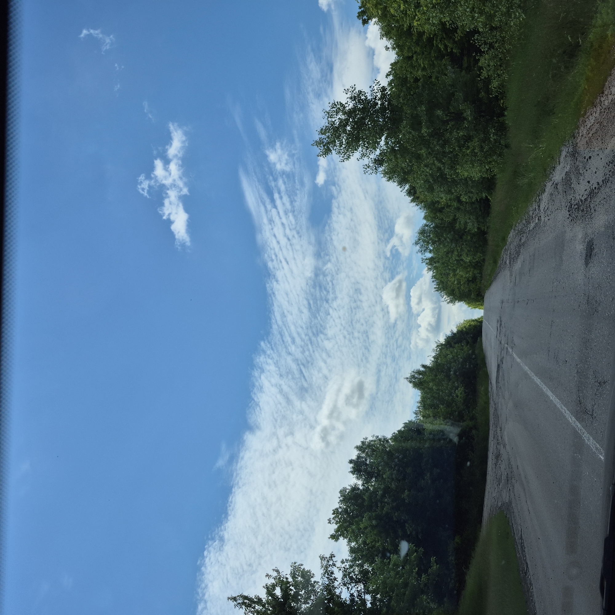 The image depicts a paved road winding through a forested area, likely in the city of Fursy, Ukraine. The road surface is worn and shows signs of disrepair, with patches of gravel and potholes. Green trees line both sides of the road, forming a dense canopy that extends into the distance. The sky above is a bright blue, with scattered white clouds, some of which are wispy and streak across the sky. The scene appears to be captured from a moving vehicle, as suggested by the perspective and the presence of what appears to be the top edge of a windshield. There are no people or animals visible in the image. The overall impression is one of a rural, somewhat neglected, but scenic road under a clear sky.