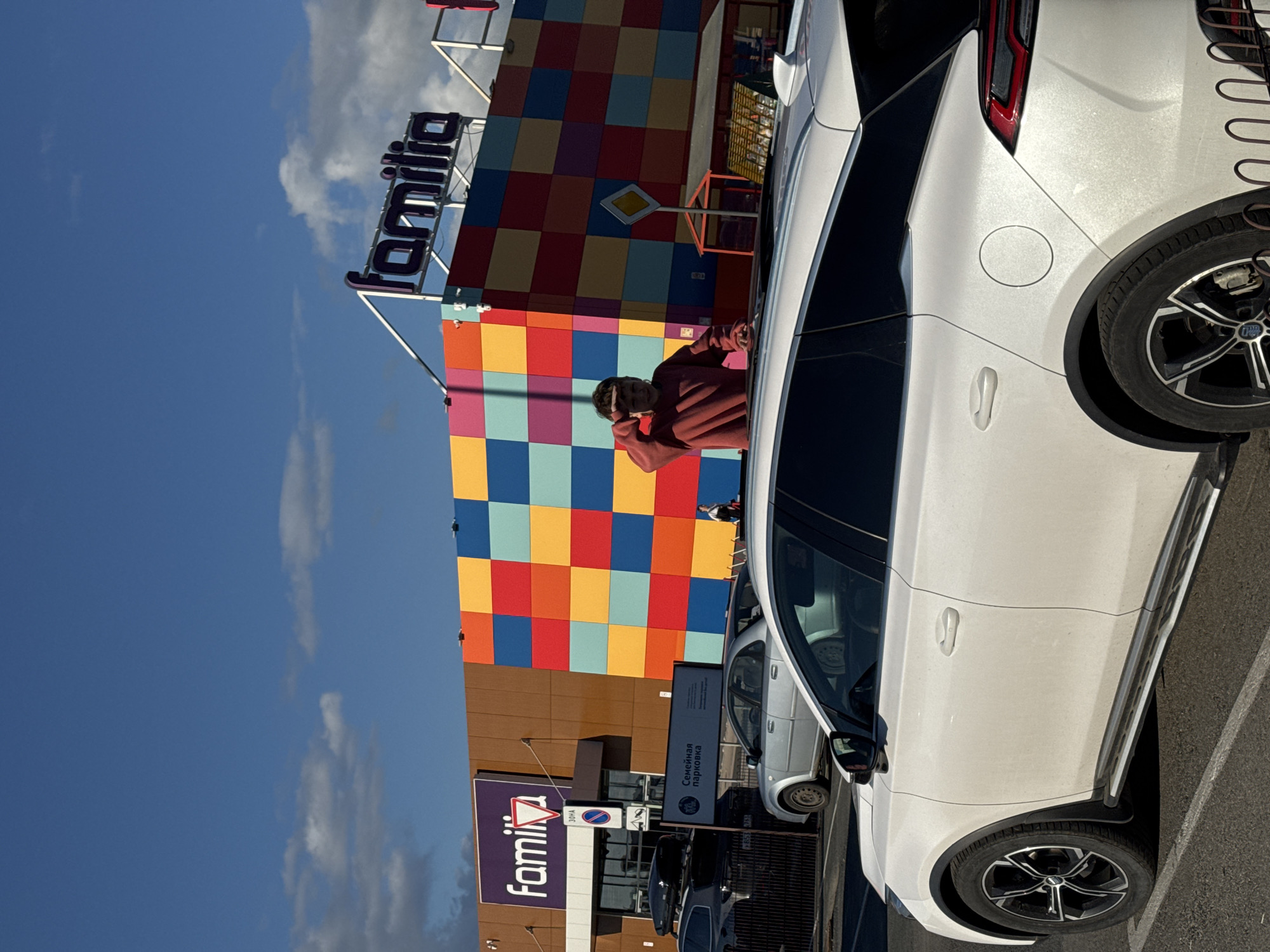 The image is a vertically oriented photograph of an outdoor scene, featuring a commercial building, parked cars, and a person under a blue sky.
On the right side of the frame, a white car, possibly an SUV or crossover, is prominently parked, appearing vertically oriented within the image. Its rear wheel, the gas cap area, and a portion of the taillight are visible. To the left of this white car, a person wearing a reddish-brown hoodie is standing. They are turned partially away from the camera, seemingly adjusting their hair or face with their hands.
Behind the person and the white car is a large building with a distinctive facade composed of bright, colorful squares in red, orange, yellow, and blue. Above this checkered section, a large purple sign with the word "Familia" is mounted. Further down on the building, a smaller sign also reads "Familia" alongside "ЗОНА" (ZONA - Zone).
Between the colorful building and the parked cars, a road sign with a yellow diamond on a white background, indicating a priority road, is visible. Another sign, a rectangular white one, has text in Russian: "Семёная парковка" (Semeynaya parkovka), which translates to "Family parking". A lighter-colored car, possibly silver, is parked further back on the left, partially obscured by the foreground car and signs.
The background shows a clear blue sky with a few scattered white clouds, suggesting a sunny daytime setting. The scene appears to be a parking lot adjacent to a large store or shopping center.