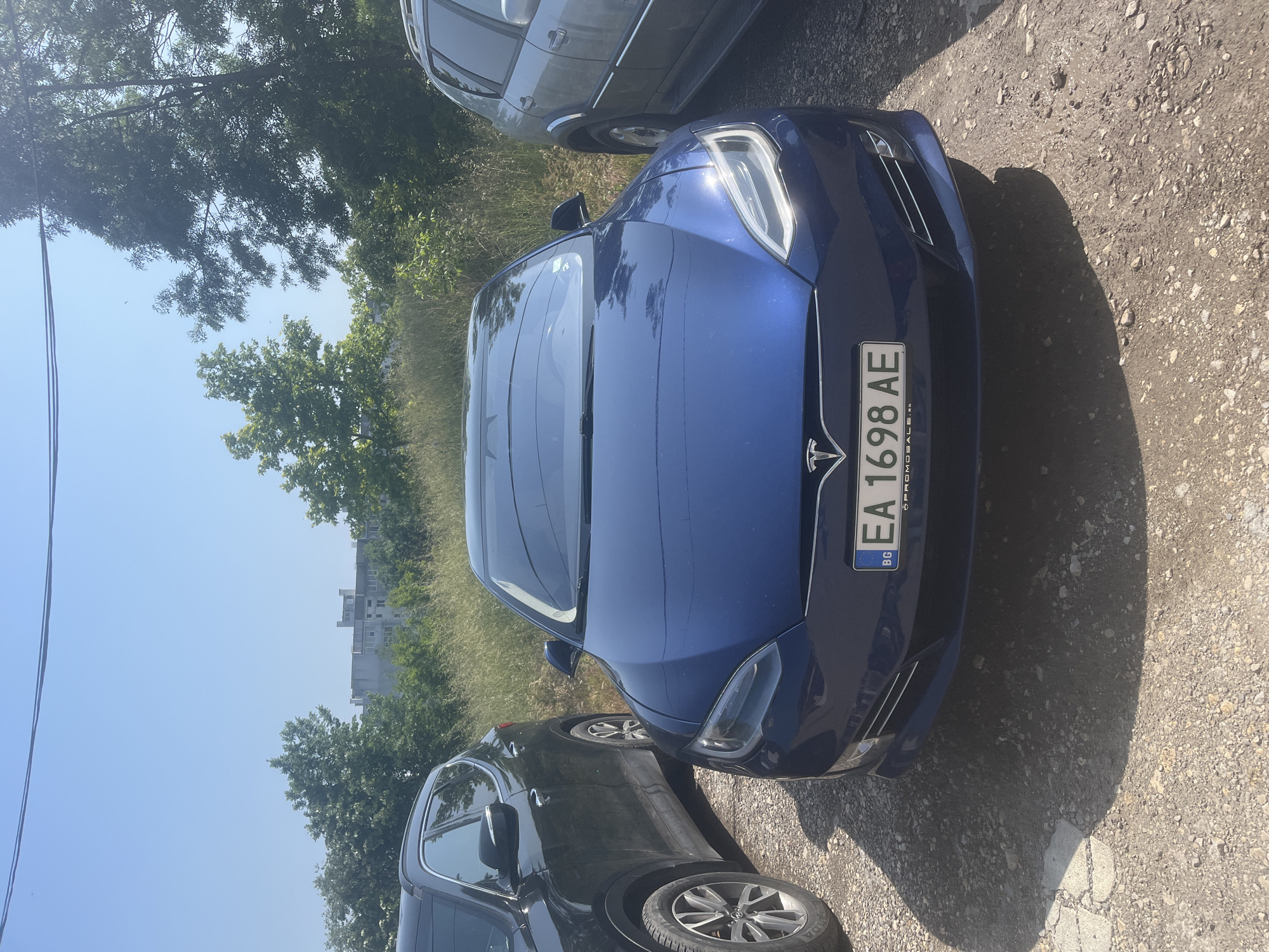 A dark blue Tesla Model S is the central subject of this image, captured from a front-facing perspective as it sits parked outdoors. The car is positioned on a coarse, gravelly and dirt surface, reflecting bright sunlight on its hood and windshield. To the left and slightly behind the Tesla, a silver or light grey car is partially visible. To the right, another dark-colored SUV, possibly a Mazda CX-5 judging by its wheel design, is also parked, with its front left wheel and part of its body in view.
The background features abundant green trees and foliage, with a multi-story, light-colored building partially visible through the trees on the left side of the frame under a clear, bright blue sky.
The time of day appears to be sunny midday or early afternoon, evidenced by the distinct, dark shadows cast by the vehicles and objects on the ground. There are no people visible in the scene, and the cars appear to be stationary.
A notable detail is the Tesla's front license plate, which prominently displays "BG EA 1698 AE". "BG" is the country code for Bulgaria. Below the main number, smaller text reads "DROMOSALE.eu". The setting is an outdoor parking area or unpaved roadside, consistent with a location in Varna, Bulgaria.