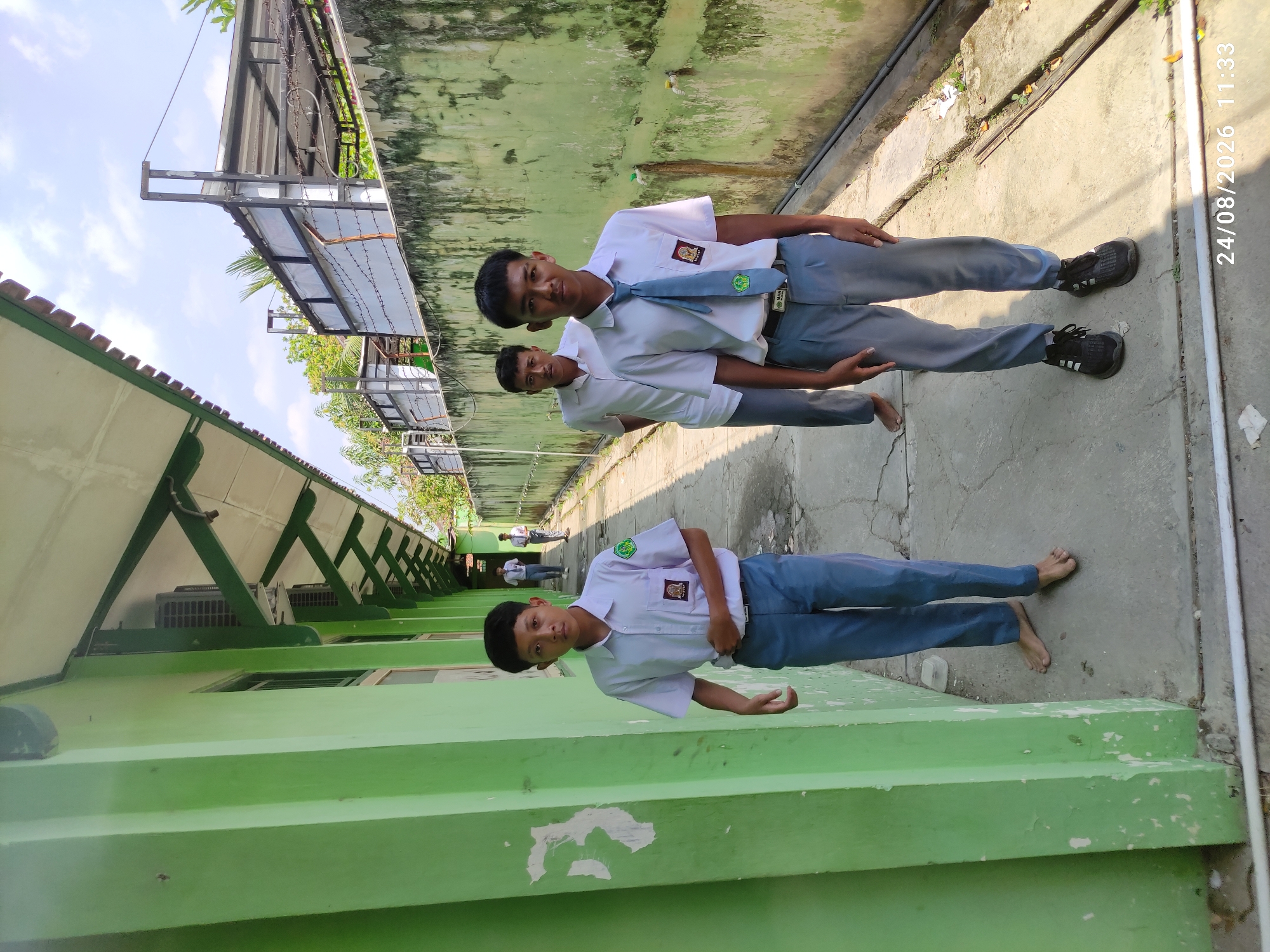 Three young Indonesian males in school uniforms stand on a paved walkway. The walkway is bordered by a bright green building on the left and a textured, weathered green wall on the right. The building has a white roof with visible rafters and air conditioning units mounted near the ceiling. The wall has several water taps and appears to be outdoors. The sky is visible above, with some clouds. The individuals are positioned at different depths, with one in the foreground facing the camera, and two others further down the walkway. The foreground subject is wearing a white short-sleeved shirt, gray pants, and black sneakers. He has a tie with a school emblem. The other two subjects are also in white shirts and gray pants, and appear to be walking or standing. The image is dated "24/08/2026 11:33". The location is specified as Tamanrejo, Indonesia.