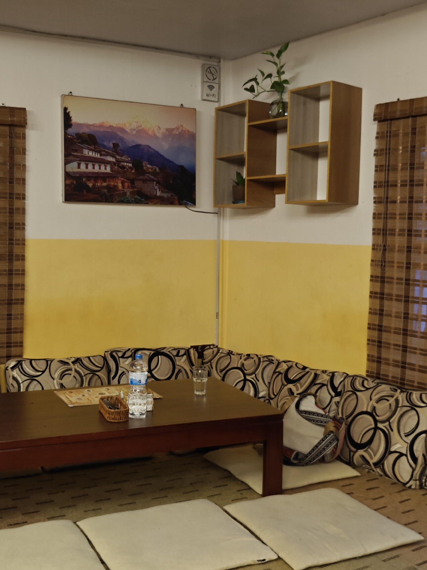The image shows a cozy seating area with a low wooden table in the foreground. The table has a few items on it, including a bottle of water, a glass, and a small wicker basket. Surrounding the table is a cushioned bench with a patterned upholstery. On the wall behind the bench, there is a painting of a village nestled in a mountainous landscape. Above the painting, there are shelves on the wall with some decorative plants. The room is lit by artificial light, and there are blinds on the windows, suggesting it is daytime. The scene appears to be in a cafe or restaurant in Pokhara, Nepal, given the artistic style and the mountainous artwork.