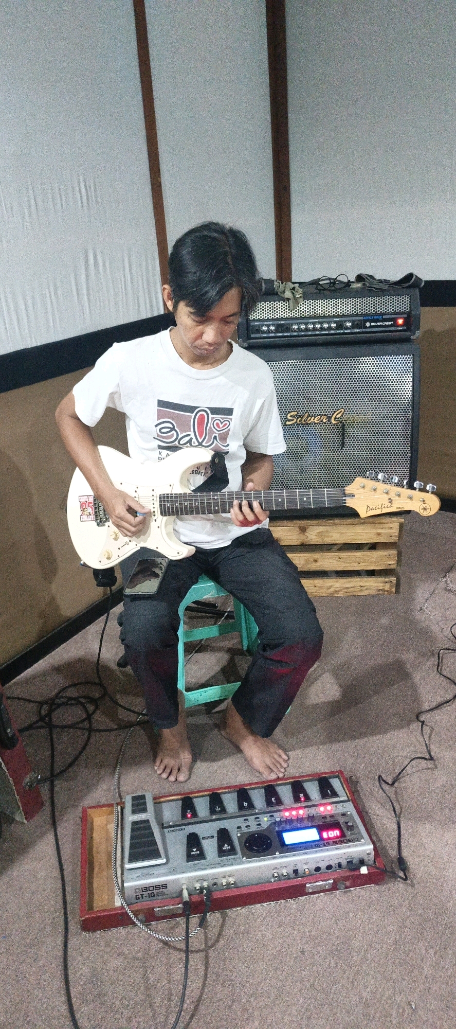 In a room with neutral-colored walls, a man sits on a teal stool playing a white electric guitar. He is wearing a white t-shirt with a graphic design and dark pants. The guitar has a "Pacifica" model designation on its headstock. In front of him on the floor is a red wooden pedalboard containing a silver BOSS GT-10 guitar effects processor. Behind the man, a two-part amplifier is visible, with a "SilverCrest" logo on the upper section. The floor is carpeted and various cables are present around the equipment. The lighting suggests an indoor setting, possibly a home studio. The scene is consistent with a music practice or recording session. The location is identified as Surabaya, Indonesia.