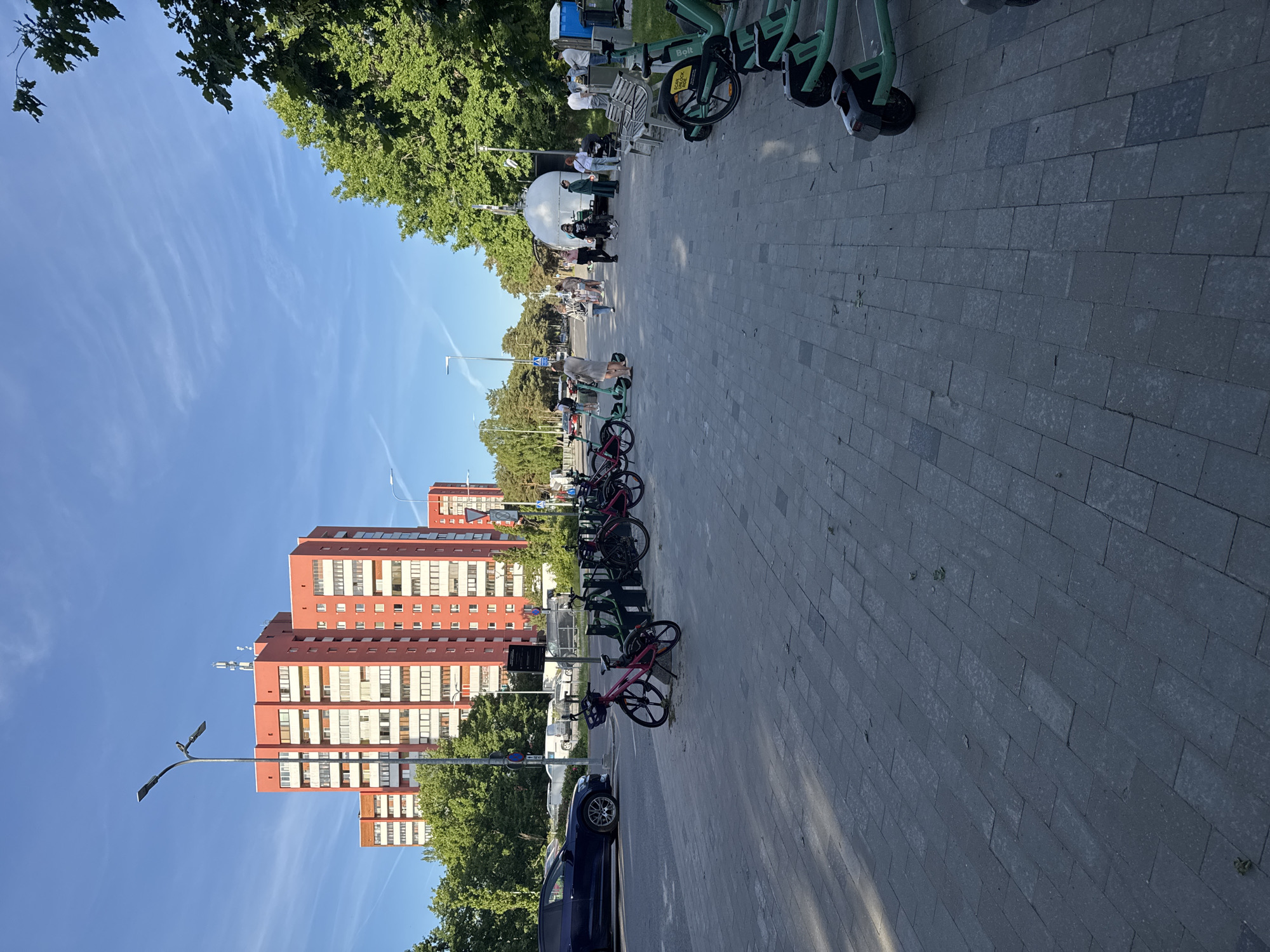 The image depicts an outdoor scene in Tallinn, Estonia, likely in a park or public area adjacent to residential buildings. In the background, several tall, modern apartment buildings with orange facades and numerous windows dominate the skyline. A bright blue sky with thin white clouds stretches above.
In the middle ground, a paved walkway is lined with rented electric scooters and bicycles. Some people are walking along the walkway, while others are gathered near a white, dome-shaped structure. A dark-colored car is parked on the street to the left of the walkway. Green trees with full foliage are visible on the right side of the image, adding to the urban park-like atmosphere. The paving stones on the walkway appear worn in places, and scattered leaves suggest it might be a warm season. The overall scene conveys a sense of everyday urban life with a focus on shared mobility options.