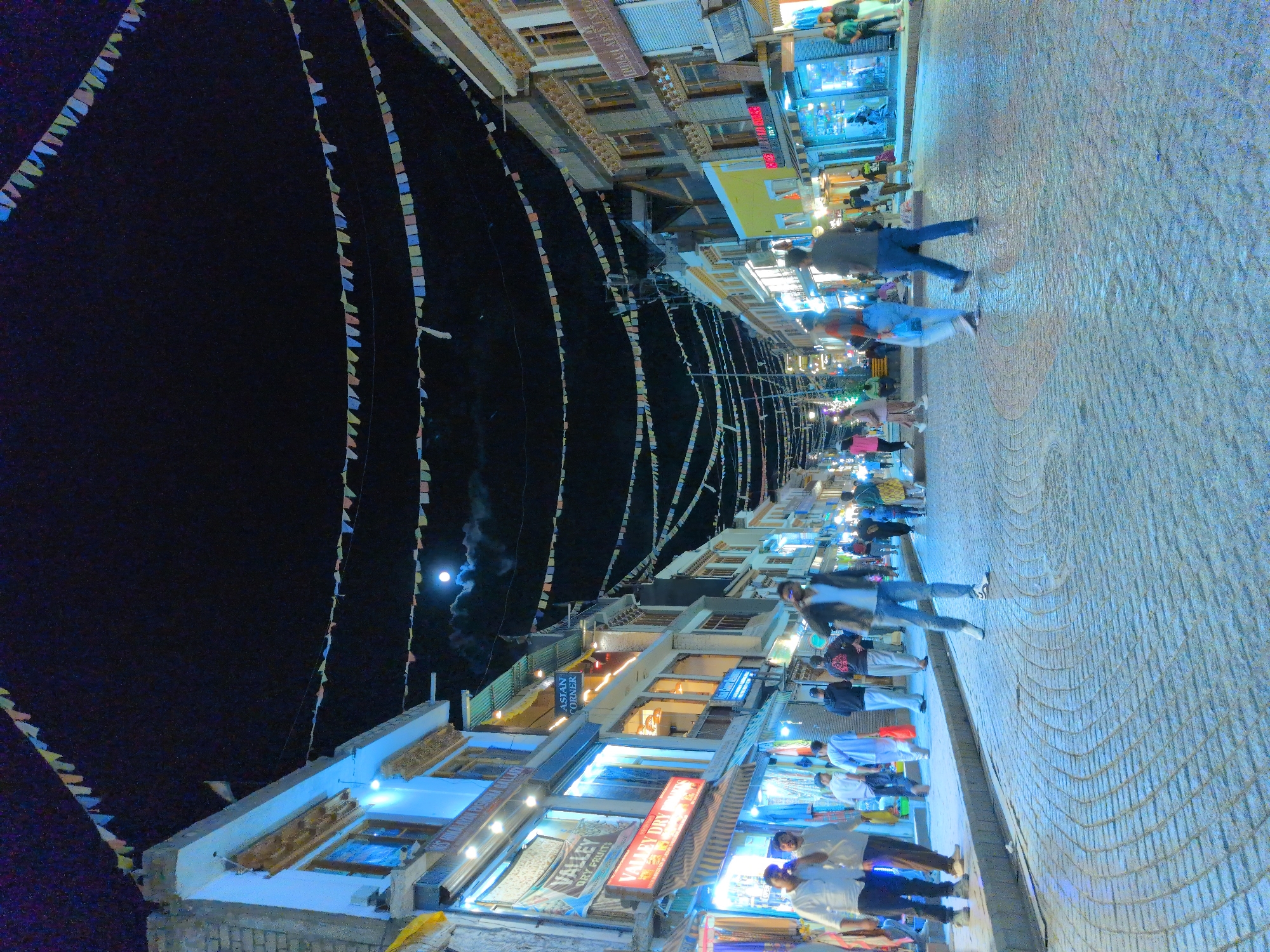 The image depicts a street scene in Leh, India, at night. The street is paved with cobblestones, and buildings line both sides, illuminated by artificial lights. Strings of colorful prayer flags are strung across the street, and in the sky, the moon is visible. Several people are walking along the street, some are shopping, and others are engaged in conversation. The overall atmosphere is lively and vibrant, suggesting a bustling market or commercial area. The architecture of the buildings, with their wooden details and layered structures, is characteristic of the region.