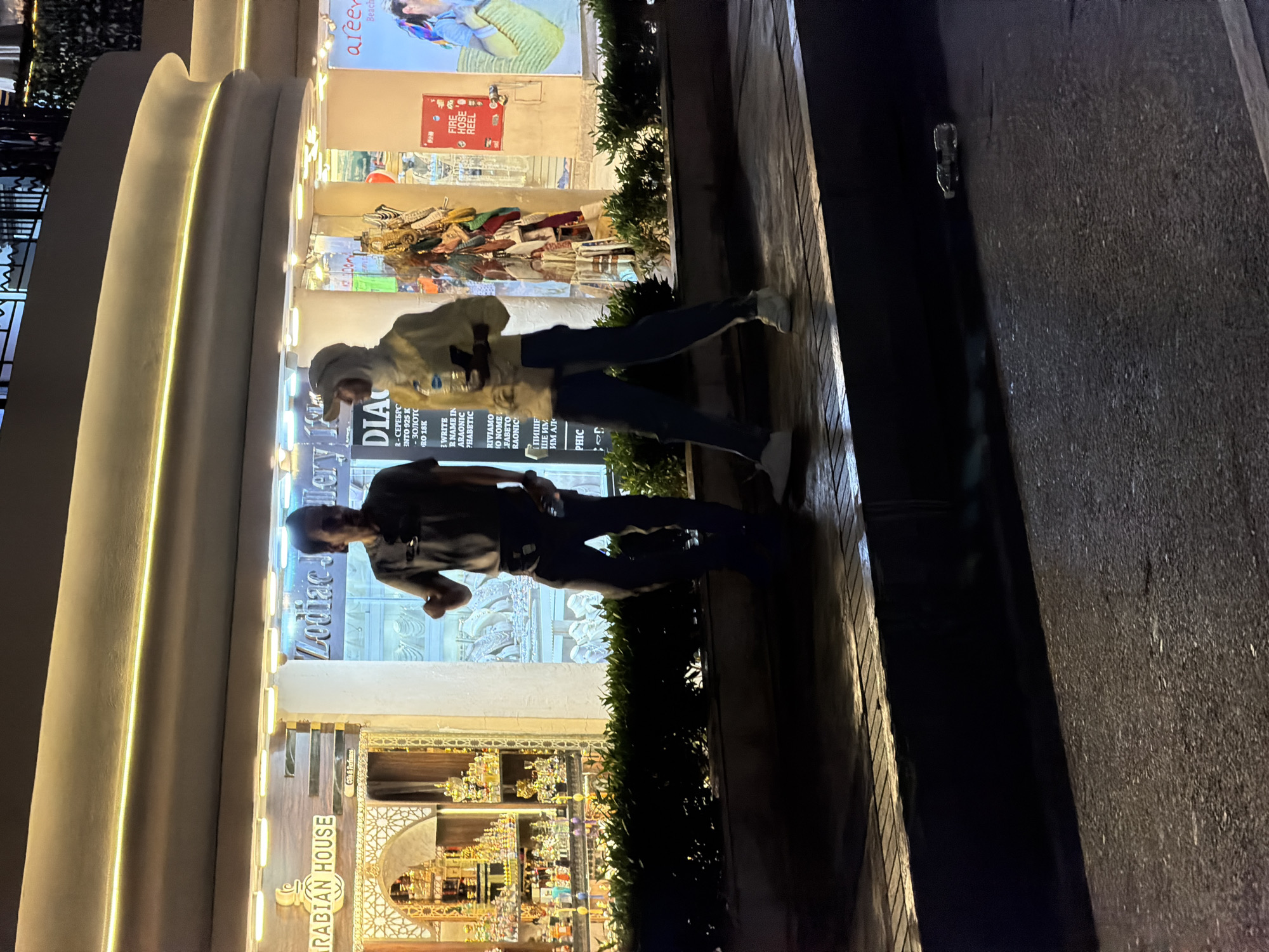 At night on a paved walkway in Sharm El Sheikh, Egypt, two individuals are walking. To the left, a man wearing a dark t-shirt and dark pants with white sneakers is seen from the waist down, facing right and gesturing with his right hand. To the right of the man, a person wearing a light-colored hooded jacket and dark pants with white sneakers is walking away from the camera. This individual is holding a plastic water bottle and a dark object in their hands. Behind the individuals are shop fronts with illuminated signs and displays. To the far left, a sign reads "ARABIAN HOUSE" with a display of small bottles. To the right of that, a sign says "Zodiac Jewlery". Further to the right, there is a display of jewelry and other items, and a red fire hose reel sign. A discarded plastic bottle lies on the ground in the lower right quadrant of the image.