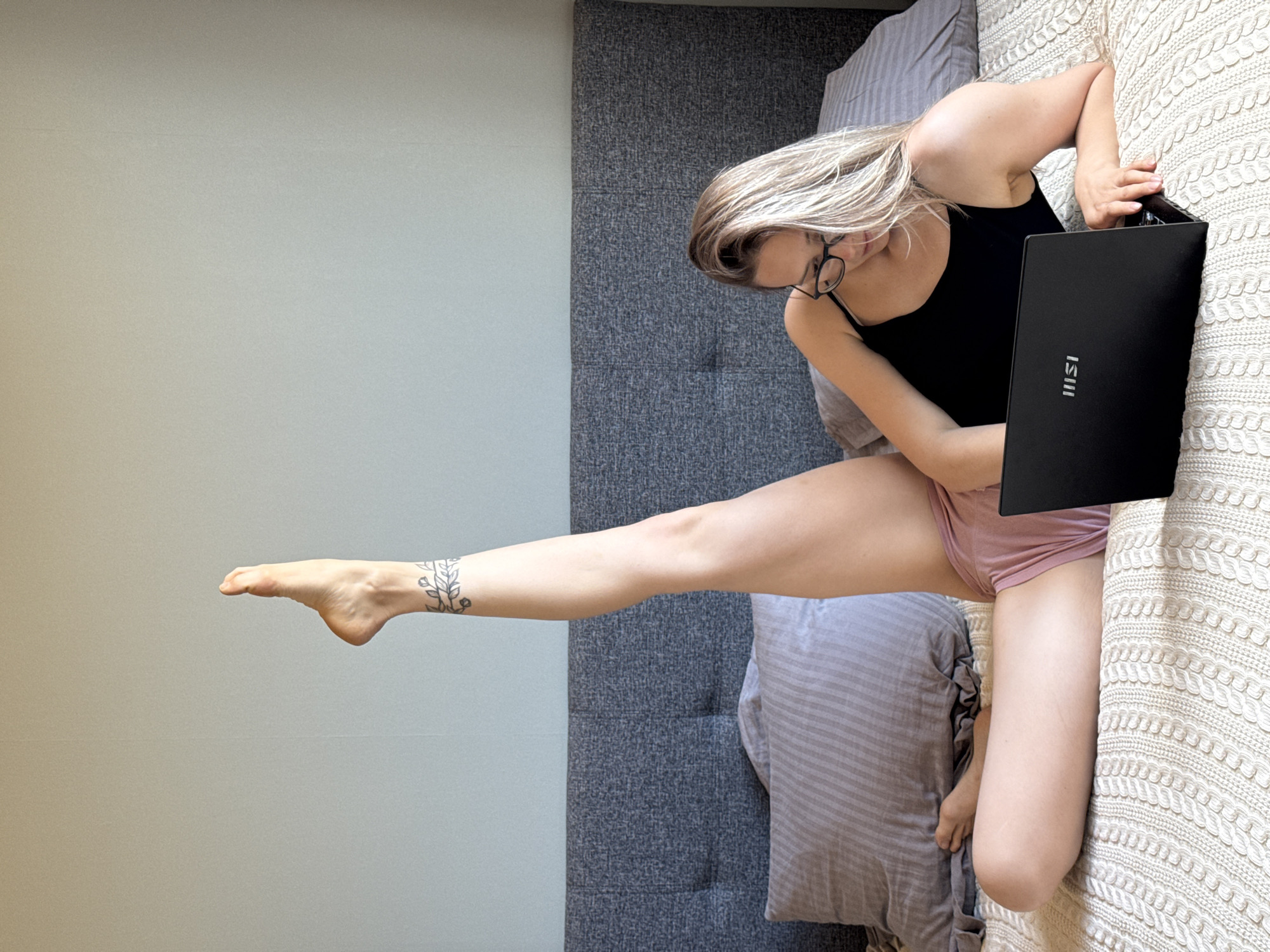 A woman with blonde hair and glasses is sitting on a bed while holding a black laptop. She is wearing a black tank top and pink shorts. Her left leg is extended towards a gray wall. There is a tattoo on her ankle. The bed has a gray headboard and a white knitted blanket. The background includes a gray wall and a gray pillow. It is impossible to determine if this photo was taken in Chaikovsky, Russia.