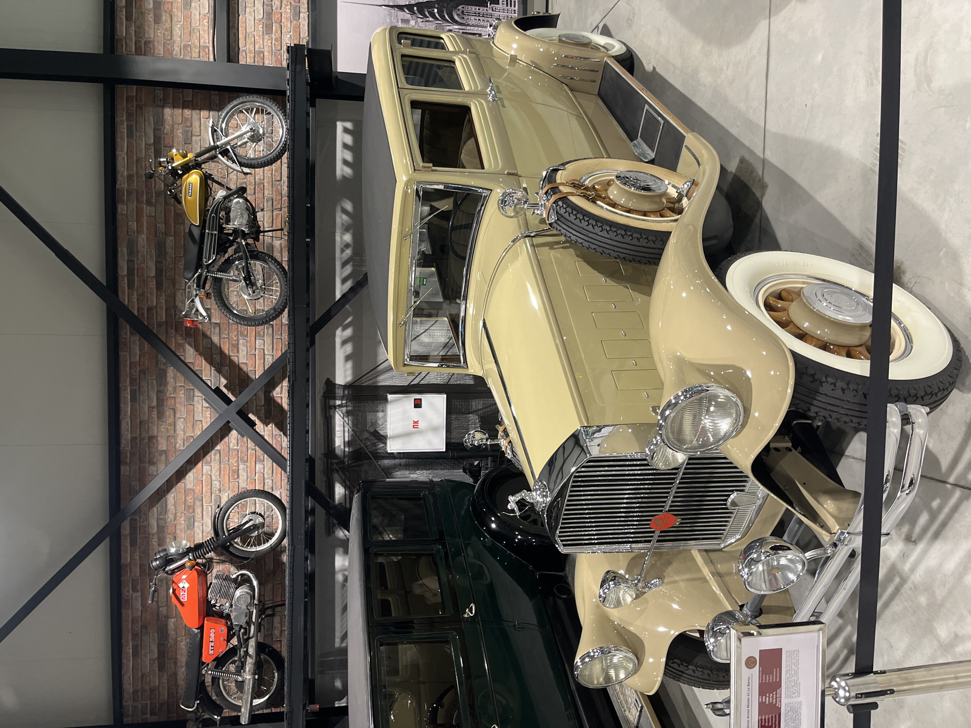 The image shows a collection of vintage vehicles inside what appears to be a museum or exhibition hall. The scene is in Kapatovo, Bulgaria.
Dominating the foreground is a beige-colored vintage automobile. It has a spare tire mounted on its side. Behind this car is another vintage automobile, colored dark green.
On the left of the frame, mounted on a brick wall, are two vintage motorcycles. The upper motorcycle is yellow and black, while the lower one is orange and black.
The automobiles and motorcycles are located on the premises in Kapatovo, Bulgaria.