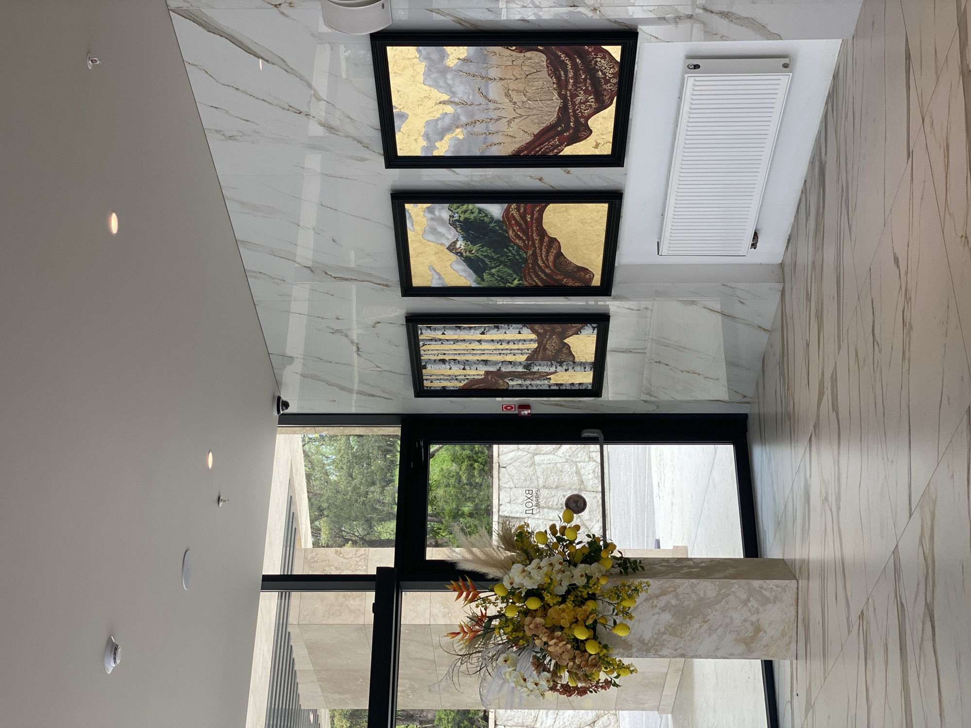 The image depicts the interior of a modern building, likely a lobby or entrance area. The walls and floors are clad in white marble with light brown veins. Three framed artworks hang on the wall, each with a black frame and depicting abstract nature scenes with gold leaf accents. Above the artworks is a white radiator.
Below the artworks is a doorway leading outside. A floral arrangement of white, yellow, and orange flowers sits on a white counter in front of the doorway. Through the doorway, one can see trees and a building exterior. The reflection in the glass door reveals the word "ЭПОХА" (EPOKHA), which is likely the name of the building or establishment.
The overall impression is one of modern elegance and understated luxury. While the image provides strong clues suggesting the building or establishment is in Sochi, Russia (such as the Russian word displayed on the doorway), definitive confirmation of the city is not directly discernible from the image alone.