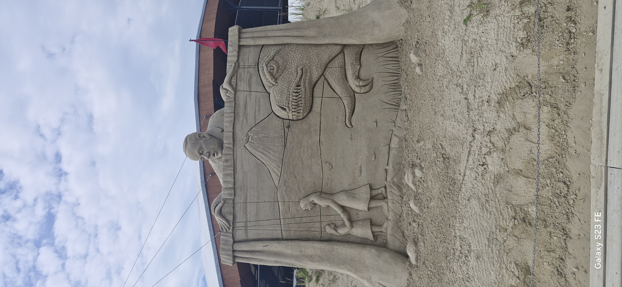 This is a photograph of a sand sculpture located on a beach, likely in Jelgava, Latvia, given the context of its creation. The sculpture depicts a puppet show scene. In the upper section, a man's head and shoulders emerge from behind a curtain, with his hands manipulating strings. Below him, a sand panel illustrates a dramatic scene. On the left, two figures of women, one adult and one child, are shown holding hands. In the background of this panel, a volcano erupts. To the right of the women, the head of a large dinosaur with sharp teeth is visible. The sculpture is rendered in detailed sand carving, with textures and features clearly defined. The environment suggests an outdoor setting, with sand, grass, and a cloudy sky visible. A red flag is planted to the right of the sculpture. The overall impression is a complex and artistic sand creation.