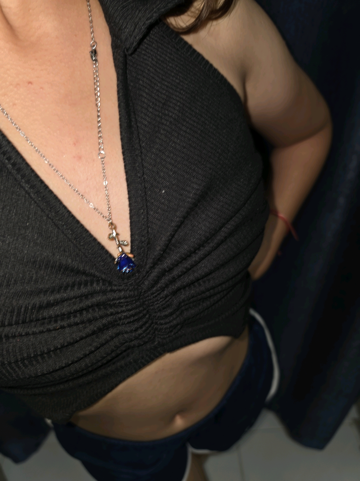 The image shows a close-up, high-angle shot of a person's torso, from the chest to the upper abdomen. The person is wearing a dark, ribbed halter top with a deep V-neckline, and dark shorts with white trim. A delicate silver necklace with a blue rose pendant hangs at the center of the chest. A thin red string is visible on the person's right arm, just above the elbow. The background appears to be a dark curtain or fabric. The scene is indoors. The image does not contain any information that would indicate a location in Phan Thiết, Vietnam.