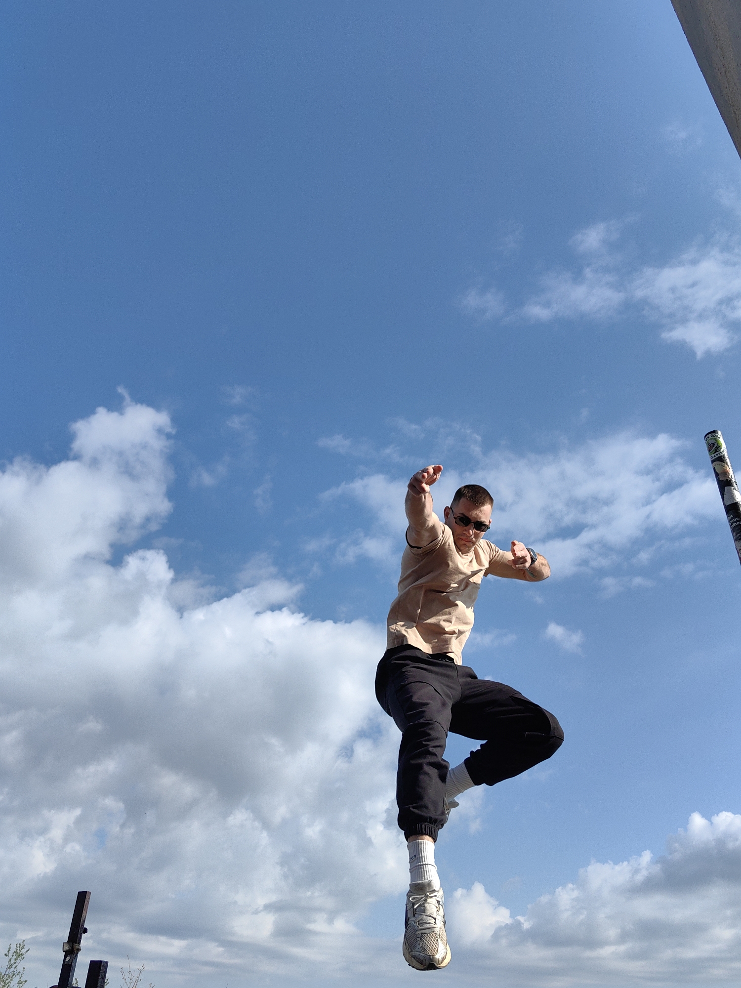 A man is mid-air, leaping, against a blue sky with scattered white clouds. He wears sunglasses, a tan t-shirt, black pants, white socks, and light-colored athletic shoes. His arms are extended, fingers pointed. A pole or pipe is visible to the right, partially obscured, and a structure appears at the bottom left. The location appears to be outdoors, possibly in an urban area. The city is Shumen, Bulgaria.