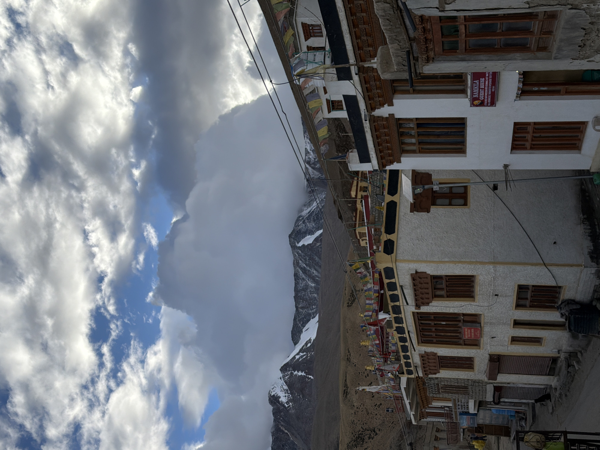 The image is an exterior shot of a street in the village of Karzok, India, with a focus on traditional architecture and the mountainous landscape. In the foreground, several multi-story buildings with white stucco facades and wooden window frames line the street. Prayer flags are strung across the rooftops of these buildings. One building on the right has a sign that reads "NAMKILA GUEST HOUSE". The street itself appears to be paved with dirt and gravel, with a black trash bin visible. Further in the background, barren, brown hills rise up to meet snow-capped mountains under a dramatic sky filled with dark, cloudy formations. The overall impression is one of a remote, high-altitude settlement with a strong cultural identity.