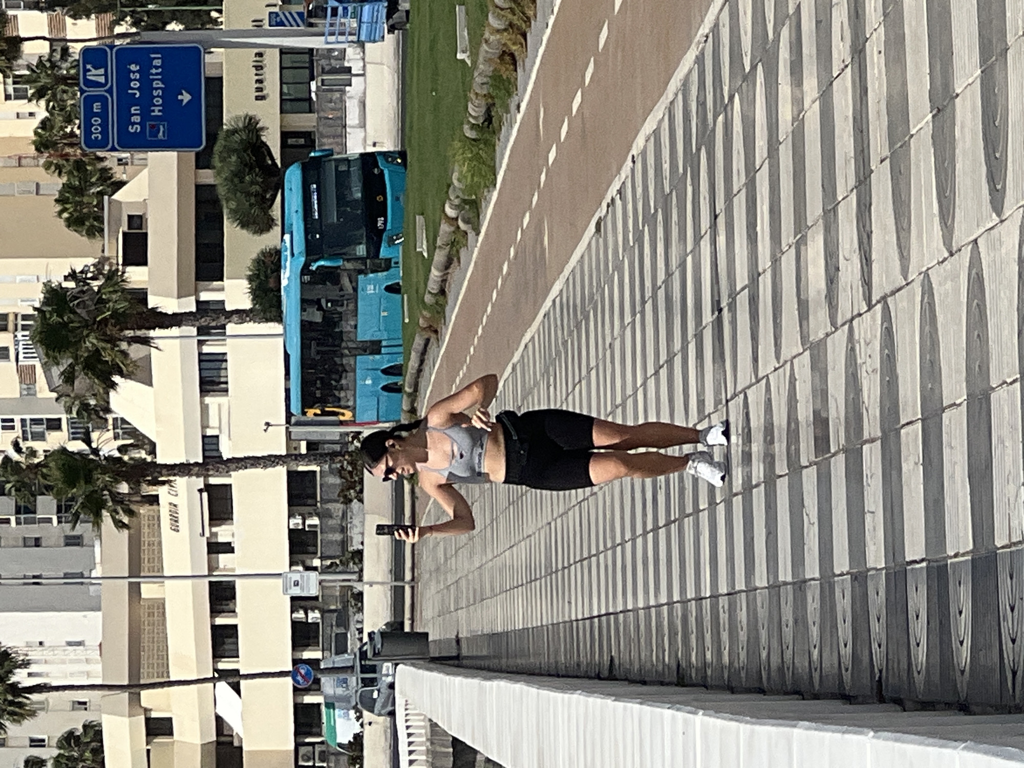 A full-length photograph depicts a woman walking along a sidewalk in what appears to be Las Palmas, Spain. She is holding a black smartphone and is wearing sunglasses, a gray sports bra, black shorts, and white sneakers. In the background, a blue bus and a building with the words "Guardia Civil" are visible, along with a blue sign indicating "San José Hospital" is 300 meters away. The sidewalk has a textured, striped pattern, and a white railing runs along its edge. A tan-colored path parallels the sidewalk, and there is grass and trees beyond that path. The surrounding area features buildings and palm trees, typical of a coastal urban setting.