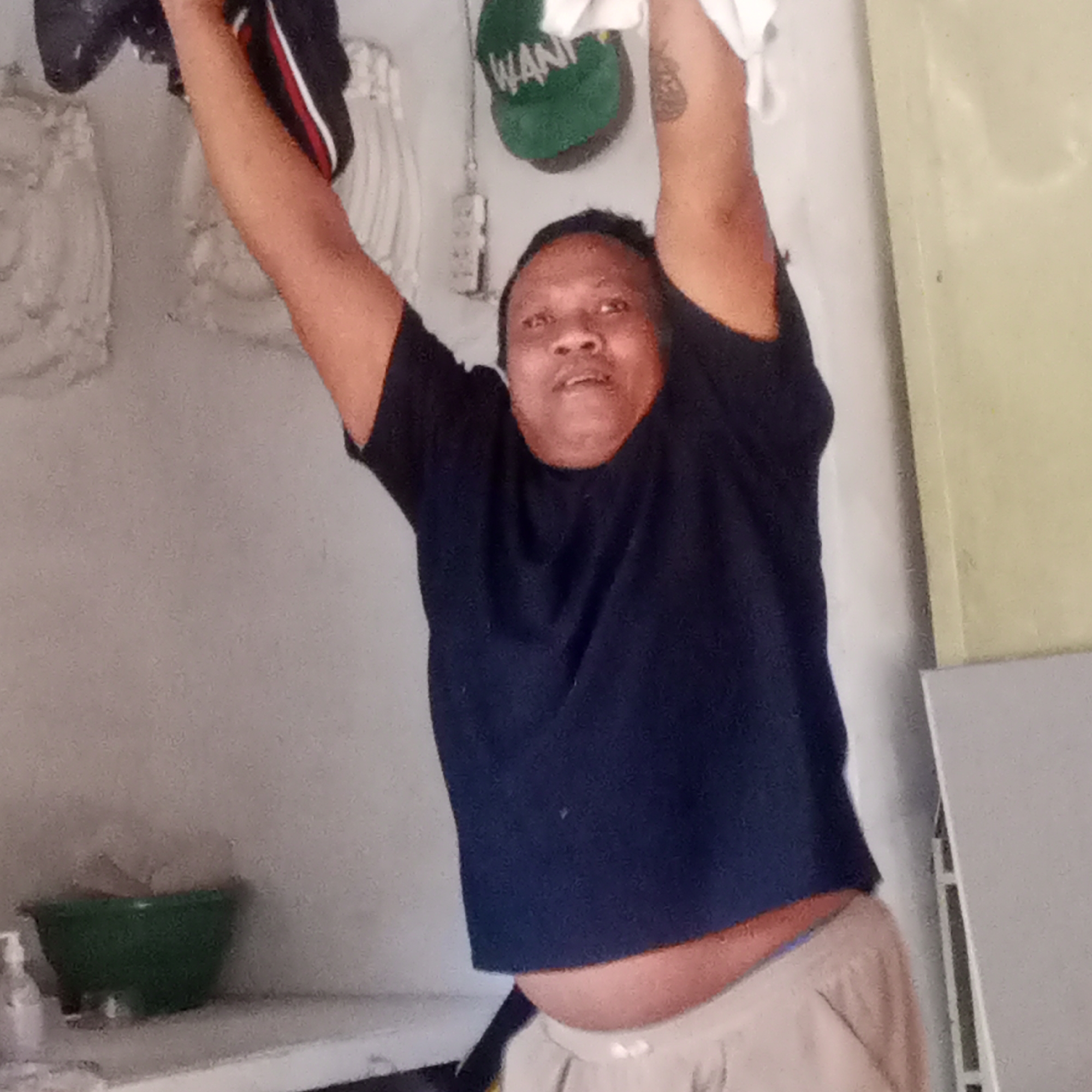 In the city of Semanding, Indonesia, a person is captured in a selfie-style photograph, their arms raised above their head. They are wearing a dark blue t-shirt and light-colored pants. The background reveals a light-colored wall and various objects, including a green bowl on a shelf. Above the person's head, a green cap with the word "WANI" is visible. The lighting suggests an indoor environment, possibly a home.
