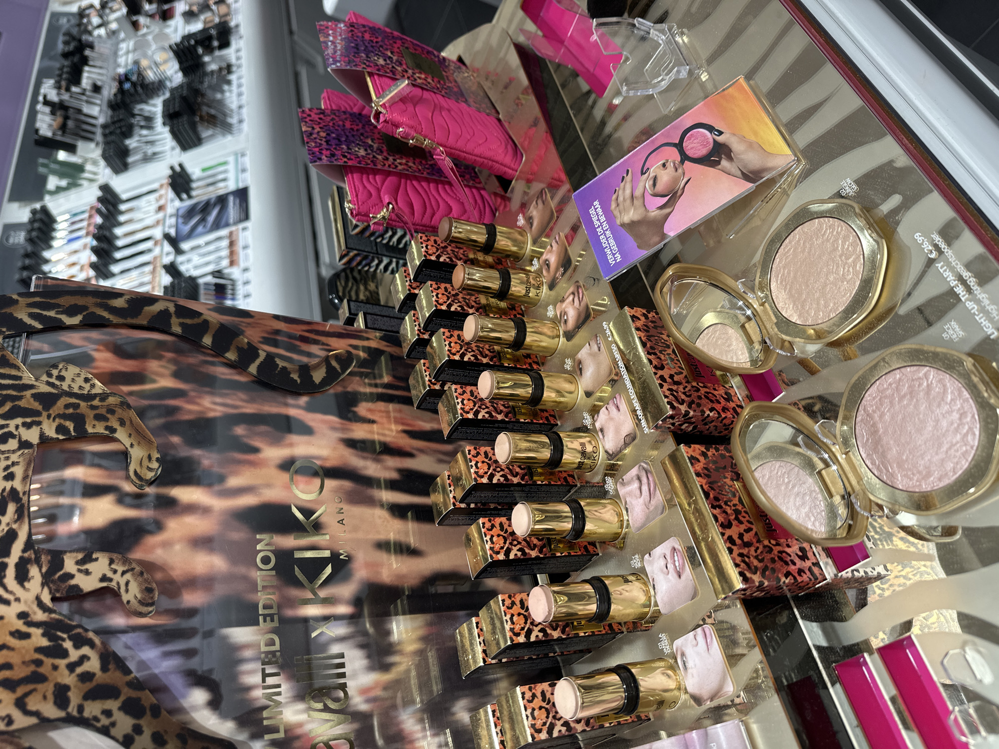 The image shows a display of Kiko Milano makeup products. Featured are sun-drape foundations, highlighters, and other items, all part of a limited edition collaboration with Just Cavalli, as noted by the branding on the display. The products are arranged on a reflective surface that shows a leopard print design. Several of the products are priced at €26.99, and the highlighters are packaged in gold-colored cases with mirrors. Other cosmetic items are visible in the blurred background, including what appear to be false eyelashes or brow products. The OCR indicates Dutch text on one of the product displays, suggesting the scene is likely within the Netherlands. No people or actions are depicted, the only action is the display of beauty products.