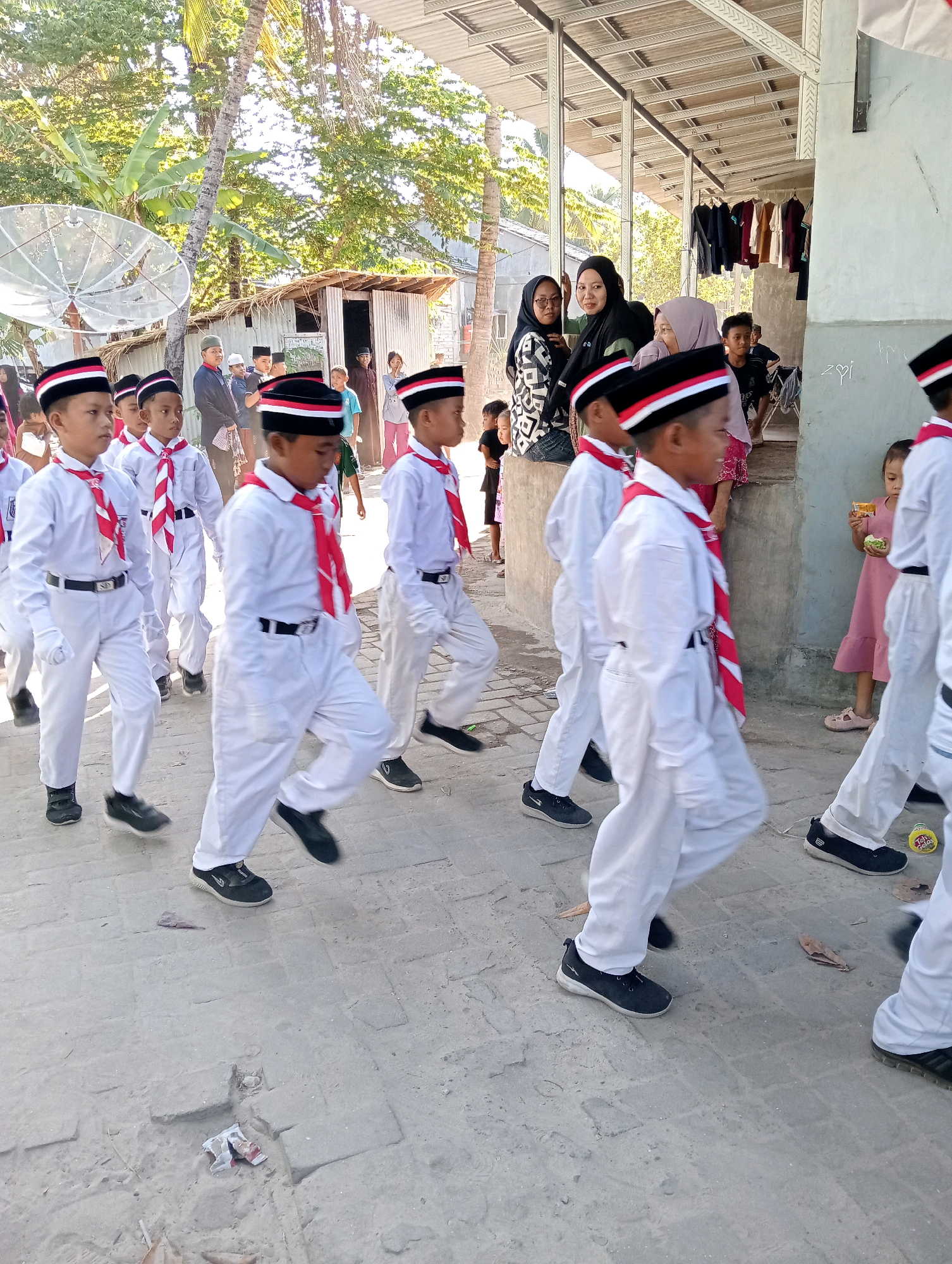 In the village of Pagerungan Kecil, Indonesia, a group of young boys in white uniforms with red and white striped ties and hats are marching in a procession. They are walking on a paved path, and in the background, there are buildings with corrugated metal roofs, palm trees, and other vegetation. Several adults are watching the procession, some are standing and some are sitting. One adult woman is wearing a hijab and patterned clothing. A young girl in a pink dress is standing to the right of the frame, holding a small object. The scene appears to be taking place during the day, with sunlight casting shadows on the ground.