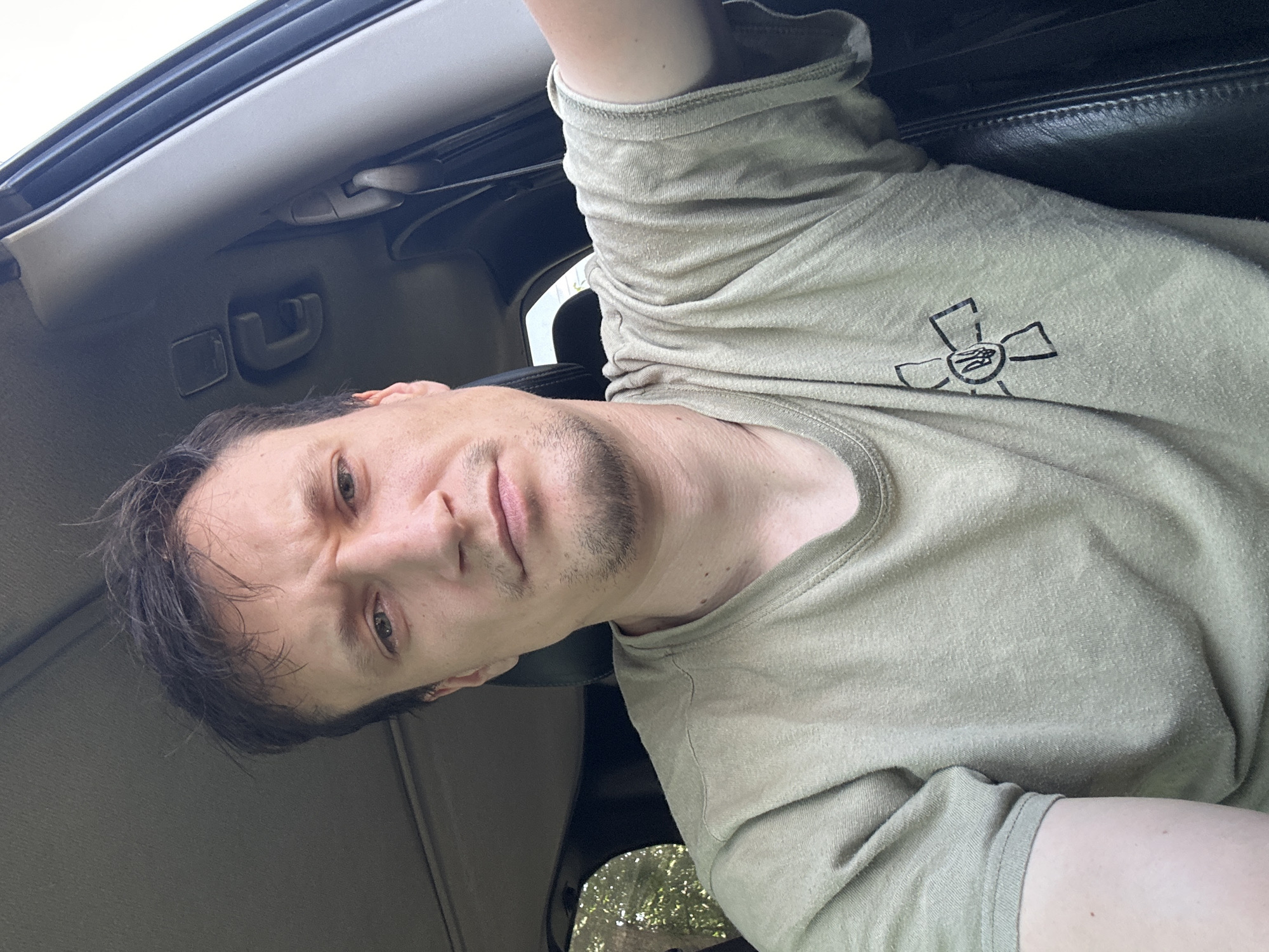 A close-up, over-the-shoulder selfie shows a man sitting in the driver's seat of a vehicle. He is wearing a light green t-shirt with a black emblem on the left breast, depicting a stylized flower with a trident in the center. The man has dark hair, a light complexion, and a slight stubble. His expression is serious, with his brow furrowed. The interior of the car is visible, including the dashboard, ceiling lining, and part of the passenger seat. Sunlight streams in through the windows, illuminating the scene. The background outside the car suggests a natural, possibly wooded environment. The image was taken in the city of Makiv, Ukraine.