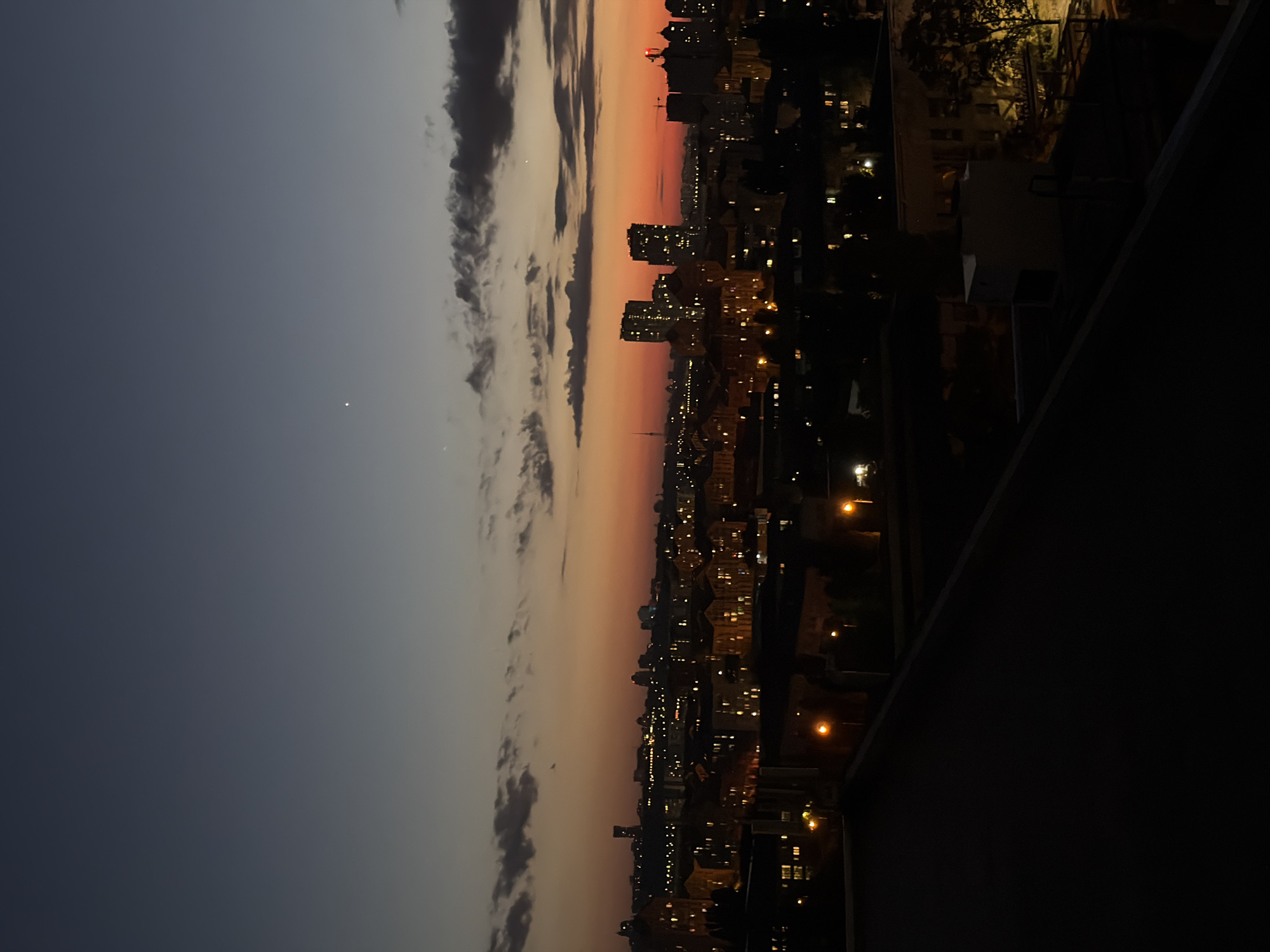 The image captures a panoramic view of Kyiv, Ukraine, at dusk. The sky transitions from a deep blue at the top to a vibrant orange and red hue near the horizon, indicating the setting sun. Several clouds are scattered across the sky, adding texture to the twilight scene. Below the sky, a cityscape unfolds, filled with buildings of varying heights, many of which are illuminated with warm, yellow lights, suggesting that it is evening and the city is coming alive with artificial light. In the distance, taller buildings and structures are visible, some with distinct architectural features, hinting at the urban landscape of Kyiv. The foreground is dominated by the dark silhouette of a rooftop, with its angular lines and dark surface creating a stark contrast to the illuminated city and colorful sky. The overall impression is one of a peaceful urban evening in Kyiv, with the city bathed in the soft glow of twilight and artificial lights.