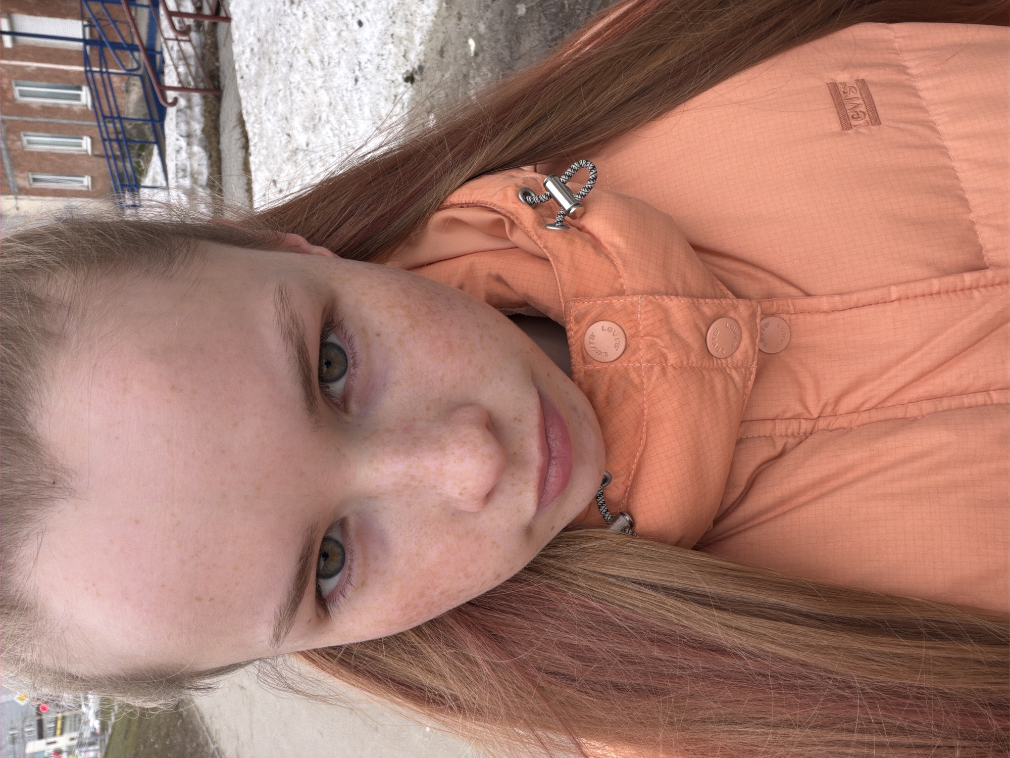 A young, fair-skinned woman with freckles and light-colored hair with reddish highlights is the focus of the image. She gazes directly at the viewer. She wears a coral-colored Levi's puffer jacket with a high collar and a small logo visible on the upper right side of the jacket.
The setting appears to be outdoors, possibly in Berezniki, Russia, based on background elements like apartment buildings with blue railings and snowy areas. These elements suggest a slightly cold climate. The composition is angled, and the background is slightly blurred, indicating a shallow depth of field.