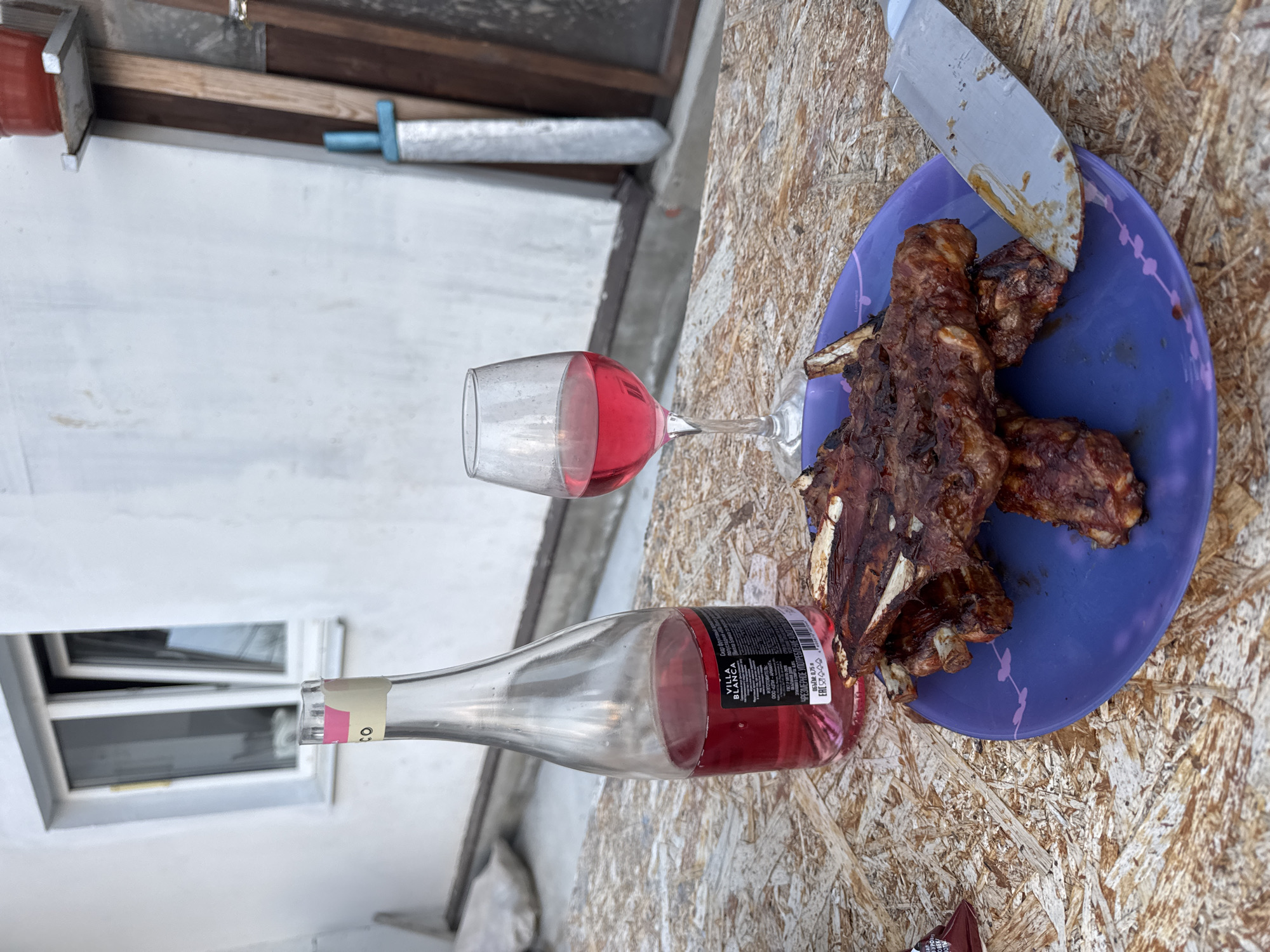 The image depicts a scene that includes food and drink. On what appears to be a wooden surface, there's a blue plate containing grilled ribs, alongside a wine glass filled with red wine, and a partially emptied bottle of "Villa Blanca" red wine. A knife rests on the plate, touching the ribs. The background includes what seems to be a window and a wall, as well as a toy sword. Based on the label of the "Villa Blanca" wine, which is manufactured by ООО «ЦЛИ - АРИАНТА», the image is potentially located in Chaikovsky, Russia.