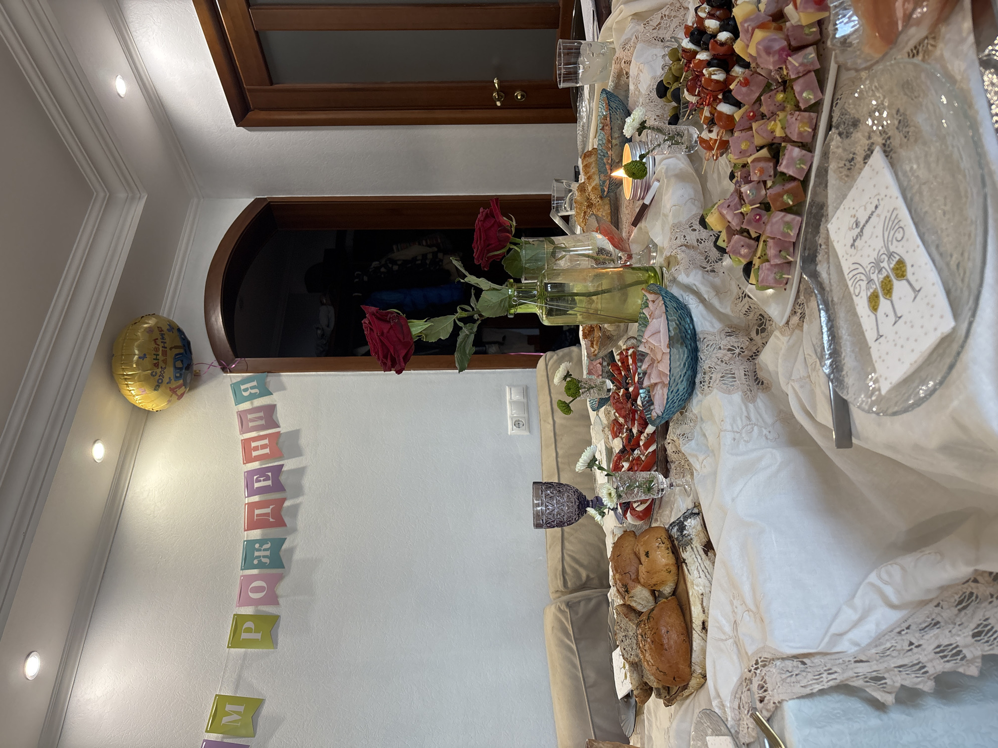 The image shows a festively decorated table inside a room. The table is covered with a white tablecloth featuring lace detailing. There are various food items on the table, including canapés, bread, and other appetizers. A vase with red roses stands on the table, along with other glassware and decorative elements. A candle is lit on the table. In the background, there is a closed wooden door and a wall decorated with a "Happy Birthday" banner in Russian (возможно, transliterated as "S Dnem Rozhdeniya"). A gold balloon is visible near the banner. The location is inferred to be Krasnoyarsk, Russia, based on the Cyrillic script on the banner. No people are visible in the image. The room appears to be well-lit and indoors.