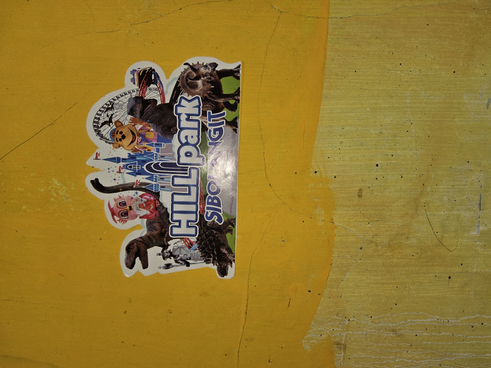 The image shows a sticker on a yellow, textured wall in Payagaboh, Indonesia. The sticker features a cartoon depiction of a theme park called "HILL PARK SIBORANGIT". The park imagery includes various cartoon characters, such as dinosaurs, a bear mascot, and a princess, along with attractions like a castle, a Ferris wheel, and a roller coaster. The wall itself shows signs of age and weathering, with cracks and dripping paint.