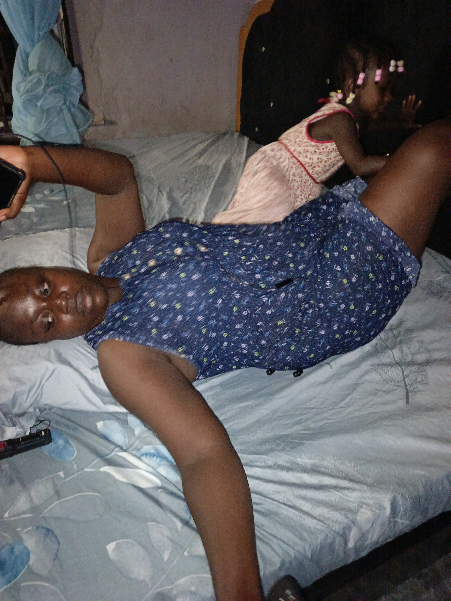 A woman is lying on a bed, with a child sitting up behind her. The woman is wearing a blue floral-patterned dress and holding a black smartphone. The child is wearing a pink dress with a red floral pattern. The bed has a blue patterned sheet. The location is likely a private residence in Akwanga, Nigeria.