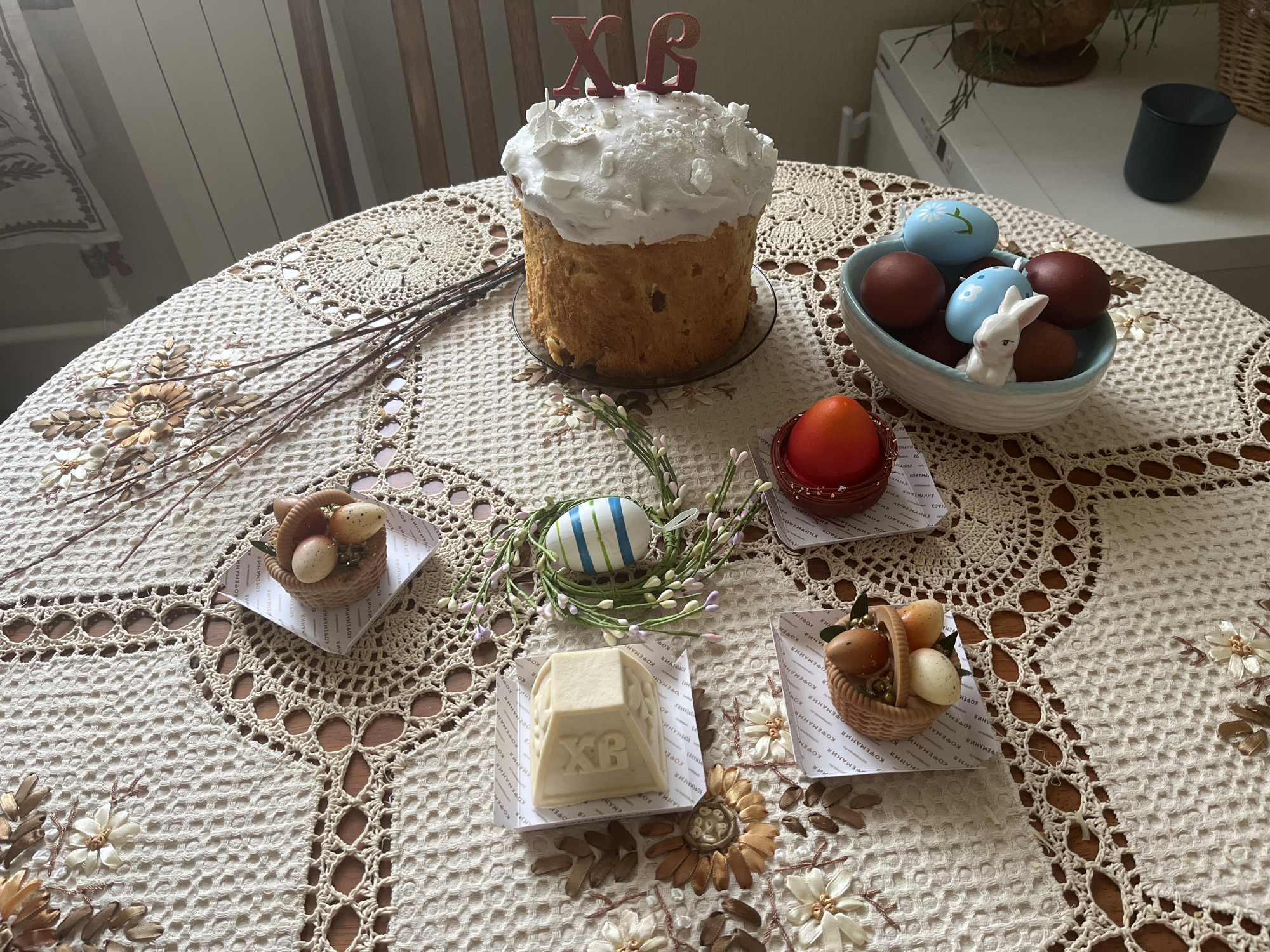 On a round table covered with a white, crochet-style tablecloth decorated with embroidered flowers, a spread of Easter treats is arranged. At the center, there is a tall, cylindrical Easter cake topped with white frosting and the letters "XB" (likely representing "Christ is Risen" in Russian). Adjacent to the cake is a light blue bowl filled with dyed eggs, predominantly brown and blue, and a small white ceramic bunny. Surrounding these focal points, there are several small, woven baskets containing decorated Easter eggs, each placed on a square of patterned paper. A white, pyramid-shaped dessert with embossed patterns sits on another square of paper. Green foliage and slender branches with buds add to the festive display. In the background, a white cabinet and a dark mug are visible, slightly blurred. The setting appears to be indoors, possibly in a home in Moscow, Russia, indicated by the text "KOPENAHHA" visible in Cyrillic on the patterned paper.
