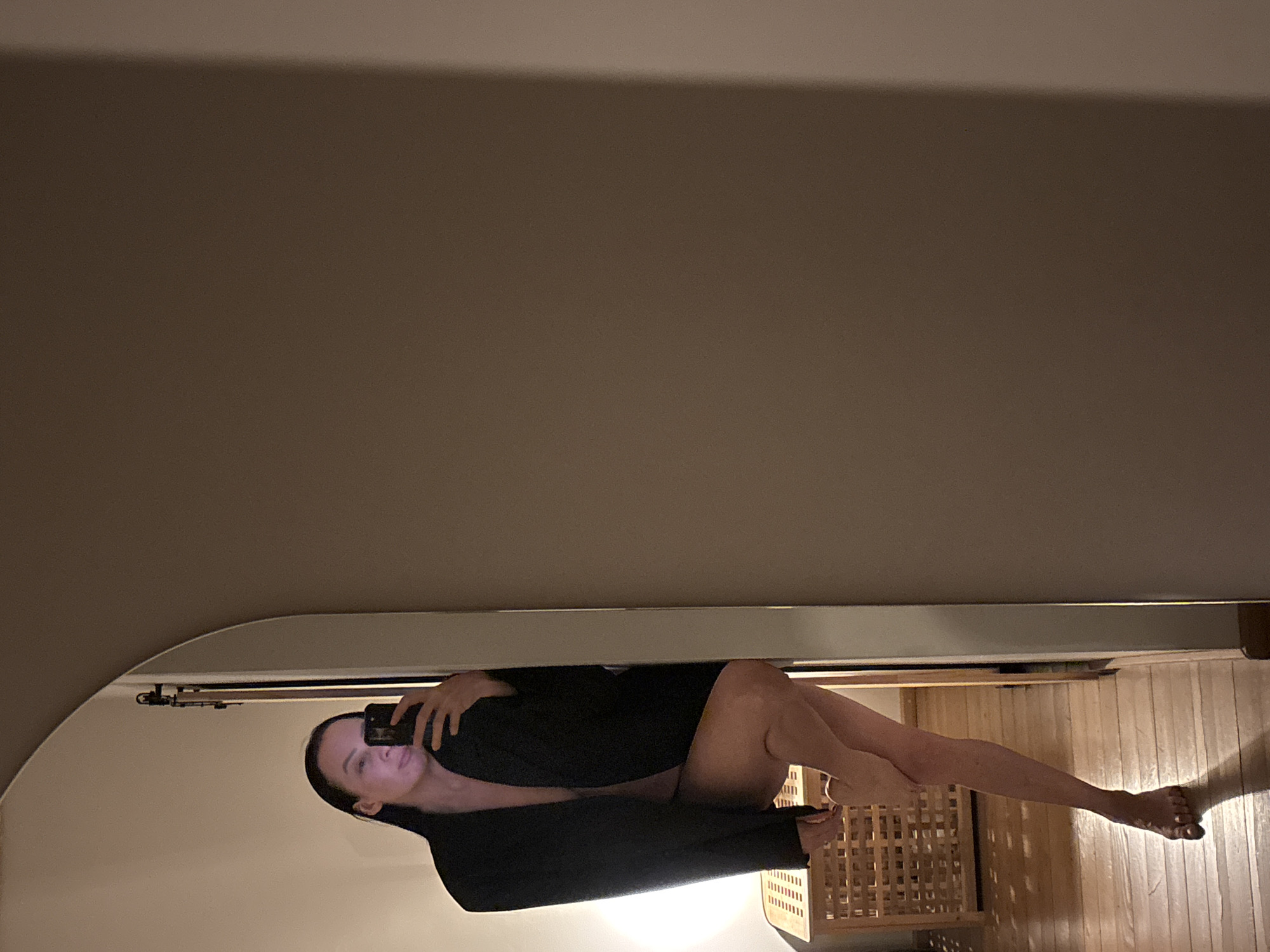 The image displays an indoor scene, a mirror selfie of an adult female. She is positioned in a tall, arched mirror, holding a dark-colored smartphone in her right hand to capture the reflection. The subject wears an open, oversized black jacket, appearing nude underneath, with her bare torso and legs visible. Her left leg is bent at the knee, while her right foot is flat on a light-colored wooden floor. Behind her lower left side, a wooden lattice-style storage unit or bench is visible. The mirror is situated within what appears to be a light-colored doorway or hallway, revealing a dark doorframe and a metal door stopper or hook in the reflection. The environment consists of light beige walls and wooden floorboards, illuminated by dim, warm artificial lighting. The specific location within the city of Chaikovsky, Russia, is not identifiable from the visual content.