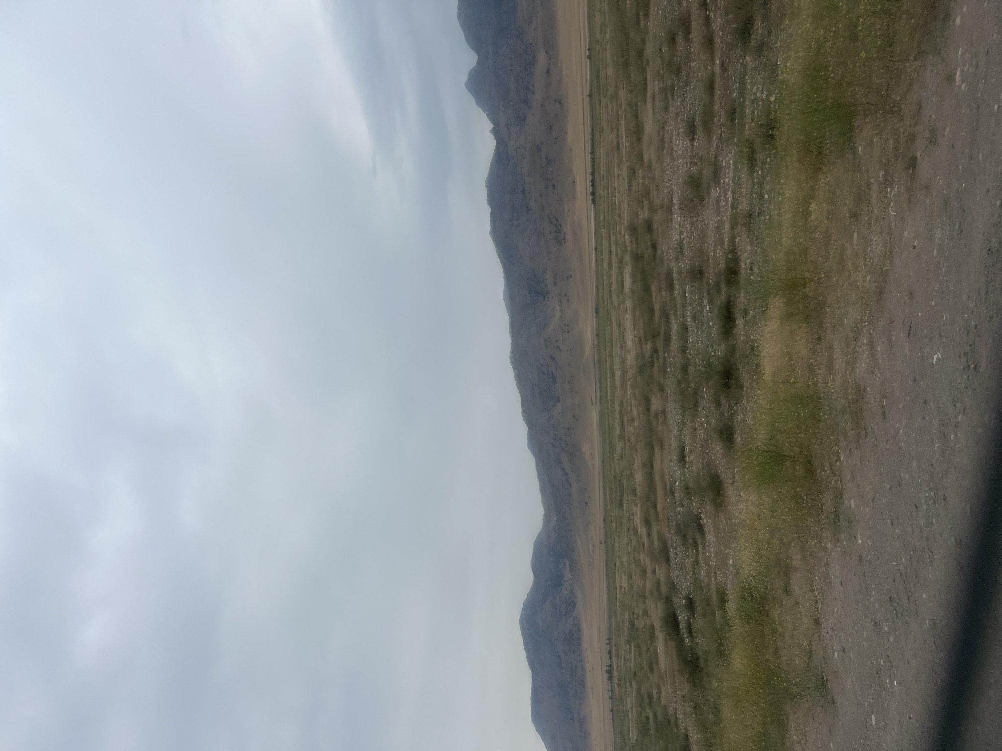 The image depicts a desolate, arid landscape under a cloudy sky. In the foreground, a dirt road or pathway runs horizontally, with dry grass and sparse vegetation dotted across its surface. Beyond the road, a wide, flat expanse stretches towards a range of undulating, barren mountains in the background. The mountains are a muted brown and grey, their slopes appearing rocky and devoid of significant vegetation. The sky is overcast with a uniform layer of grey clouds, suggesting either an impending storm or a generally dim day. The scene conveys a sense of remoteness and a harsh, dry environment. The location is identified as Chim-Korgon, Kyrgyzstan. There are no people or animals visible in the image, nor any structures or signs of human activity beyond the dirt path.