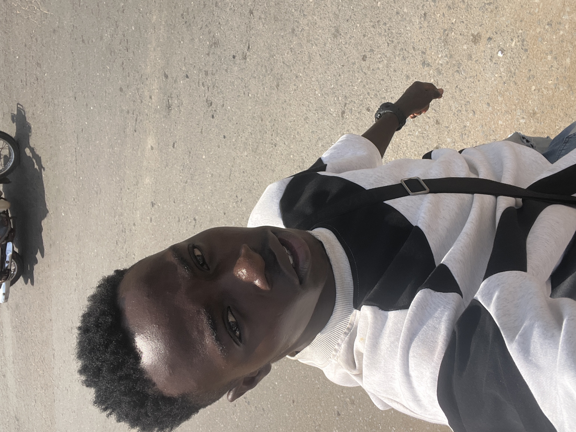 This image, taken in Onitsha, Nigeria, features a young Black man posing outdoors during the daytime. He is positioned against a light grey, textured paved surface, likely a road or sidewalk, with his head on the right side of the frame as if the image were rotated.
The man has a dark complexion, short, tightly curled black hair, and dark eyes. His face appears slightly shiny, possibly from the bright sun or natural oils. He wears a distinct white and black color-blocked long-sleeved shirt or sweatshirt, with a black strap, likely from a bag, draped across his chest. A dark watch or wristband is visible on his left wrist. Partial glimpses of light blue jeans can be seen.
The background shows the continuation of the paved ground. To the left, a clear shadow of a motorcycle or scooter is visible, along with a small part of its front wheel and some metallic components. The strong, direct lighting suggests it is a sunny day, casting defined shadows. There is no visible text in the image.