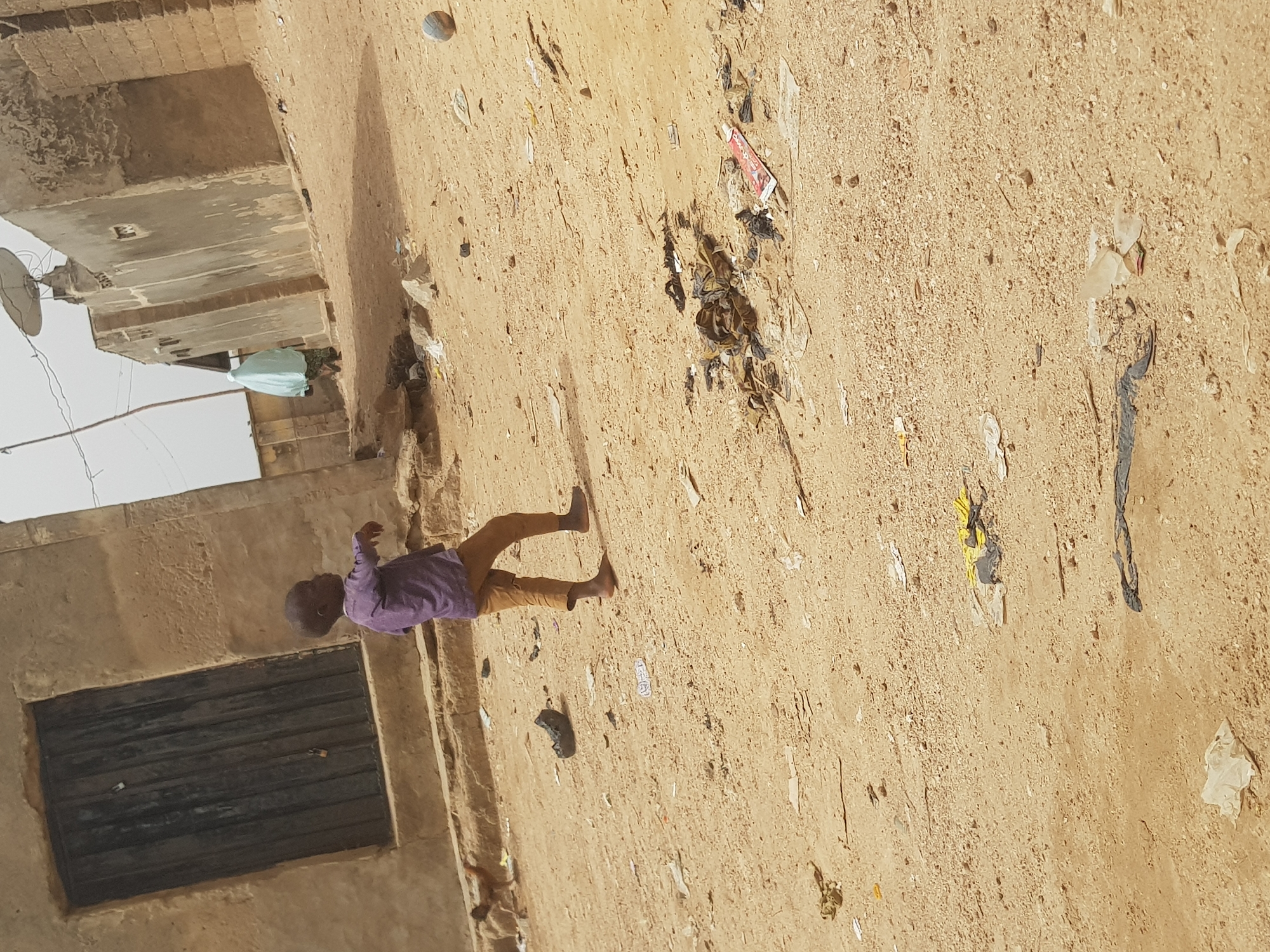 A daytime shot shows a child walking barefoot on a dirt ground in Dutsinma B, Nigeria. Litter is scattered on the ground. The child is wearing a purple shirt and brown pants. To the left is an old building with closed, barred windows. A satellite dish is mounted on the building's roof.