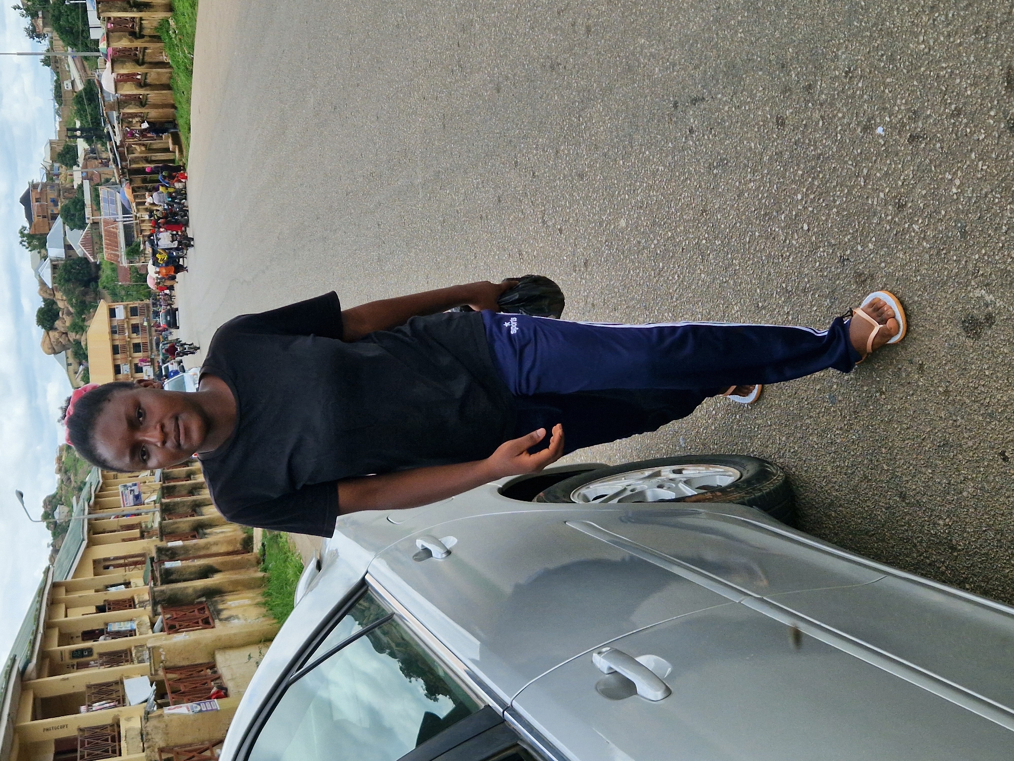 A woman walks on a paved road beside a silver car. The woman is wearing a black t-shirt and blue track pants. She is holding a black plastic bag in her right hand. In the background, there are buildings, people, and motorcycles. The sky is overcast.