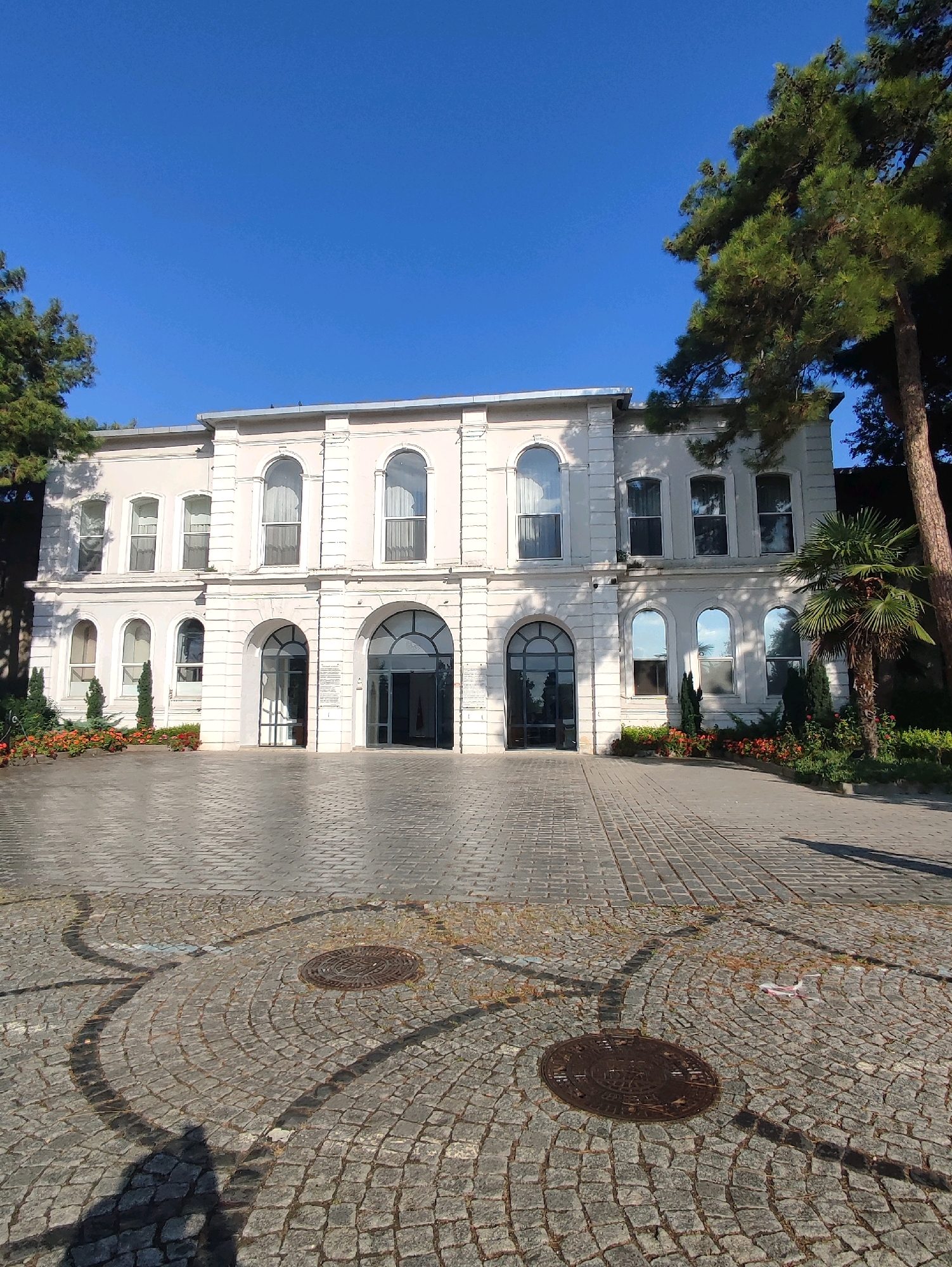 This image depicts a two-story building with a light-colored facade and arched windows. The building is situated in front of a clear blue sky and surrounded by greenery, including trees and shrubs. The foreground is paved with cobblestones, arranged in a circular pattern around two manhole covers. The scene is illuminated by natural light, casting shadows on the ground. There are no people visible in the image. The architectural style suggests it could be a historical or institutional building, possibly located in a park or a well-maintained outdoor area. The image is taken from a low angle, emphasizing the building and the surrounding landscape.