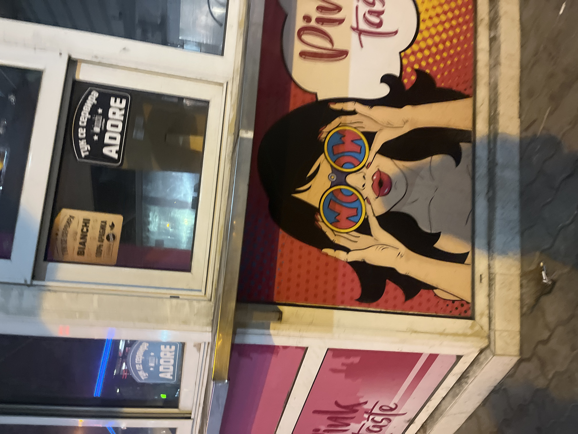 A medium shot depicts a storefront in Sofia, Bulgaria, specifically an establishment called "Pink Taste," as indicated by signage. The sign features a stylized cartoon image of a woman with black hair, wearing round sunglasses with the word "WOW" imprinted on the lenses. Her red lips are visible, and she is positioned with her hands near her face in a gesturing pose. The storefront also has a separate sign that reads "ADORE." The environment is urban, as suggested by the street pavement visible on the right and the appearance of reflective surfaces in the storefront windows.