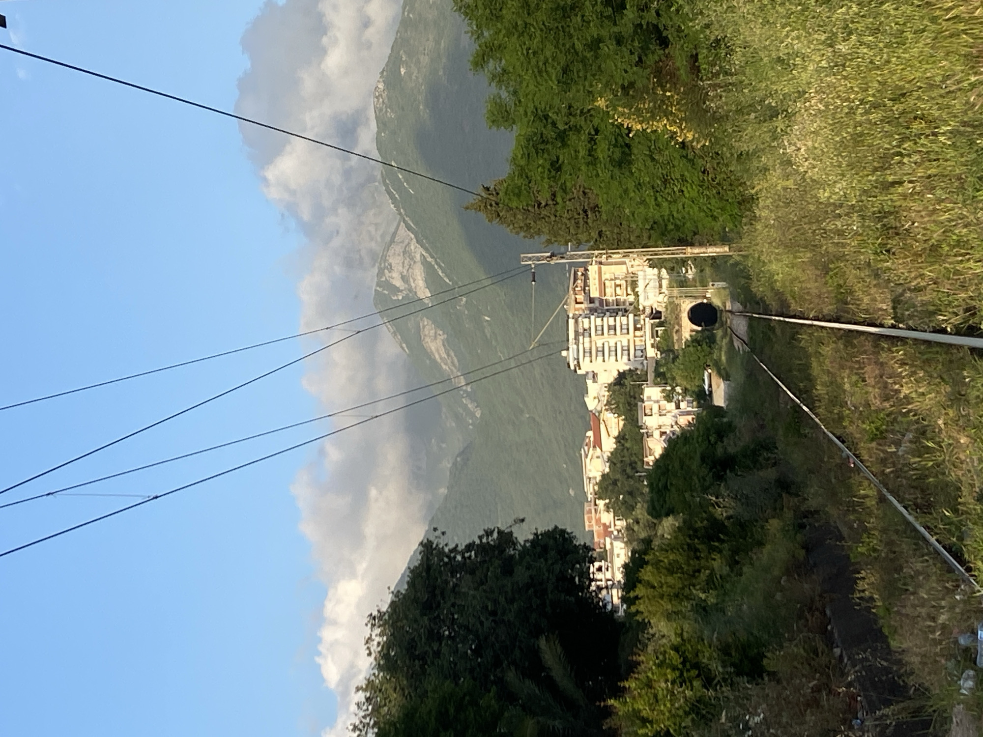 The image shows a railway track in Bar, Montenegro. The track leads to a tunnel. There are buildings on the left side of the tracks, including a multi-story white building. A mountain is visible in the background, partially obscured by clouds. Trees and greenery line both sides of the railway track. There are power lines overhead.